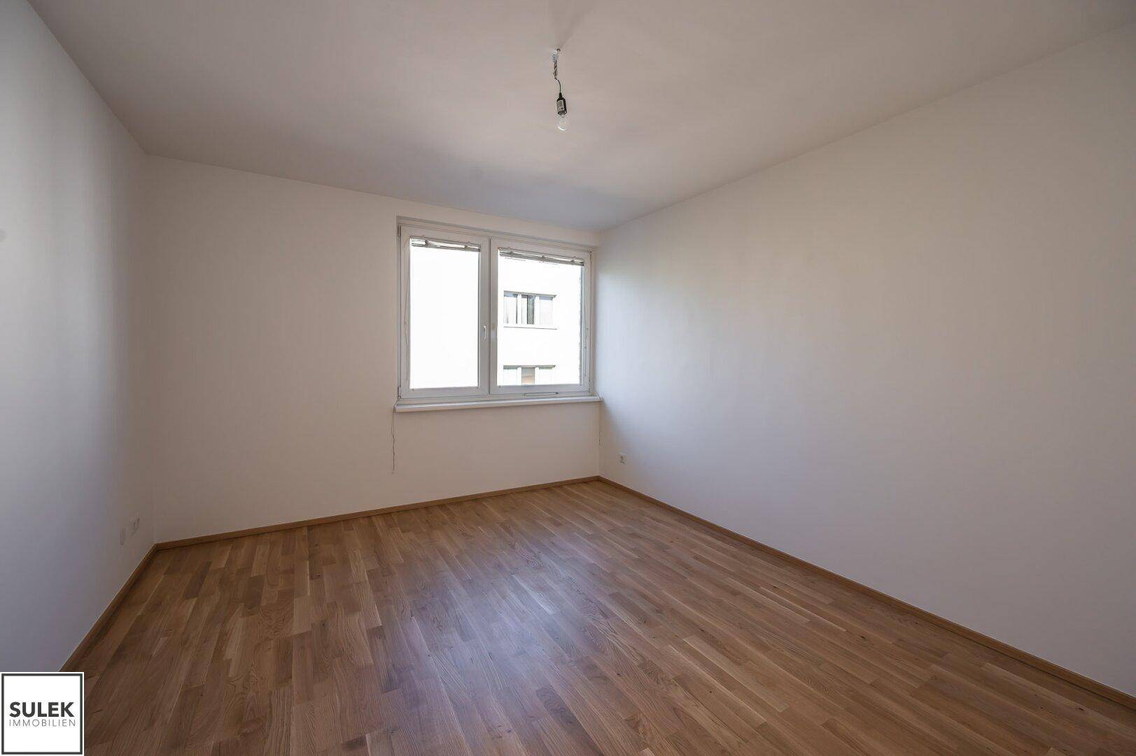 Geräumiges, leeres Zimmer mit Parkettboden und einem großen Fenster, das den Raum erhellt.