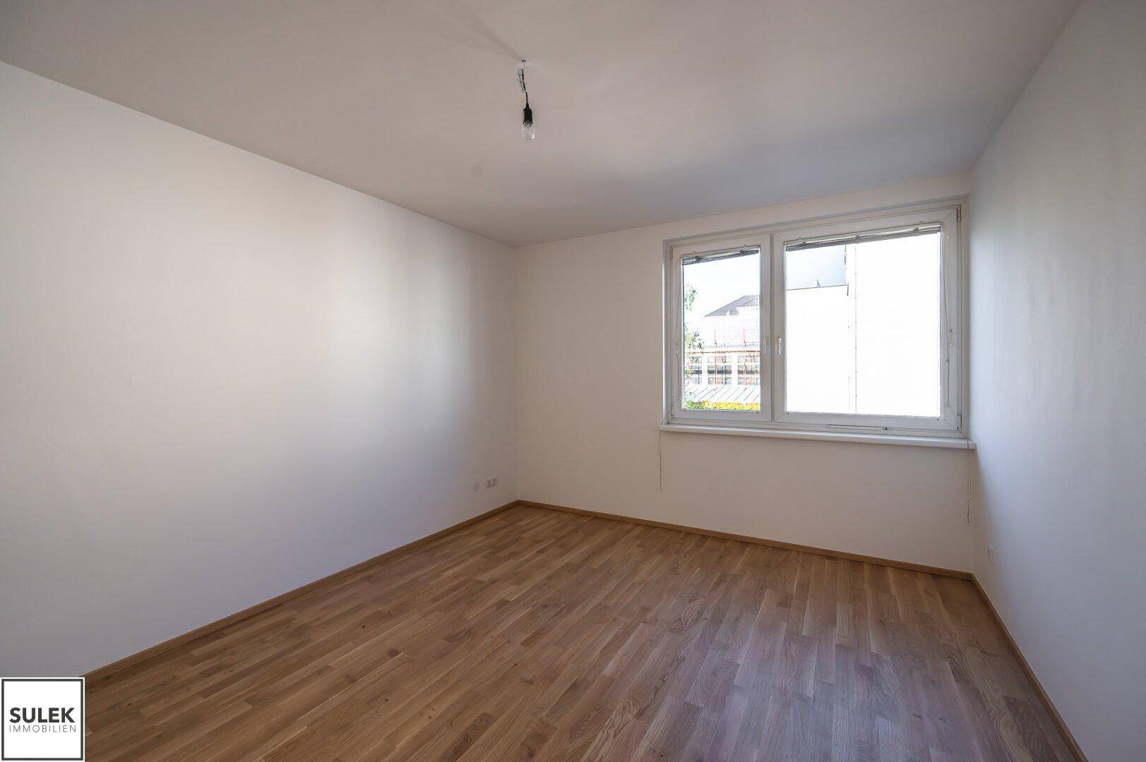 Leeres Schlafzimmer mit Parkettboden und einem Fenster, das viel Tageslicht hereinlässt.
