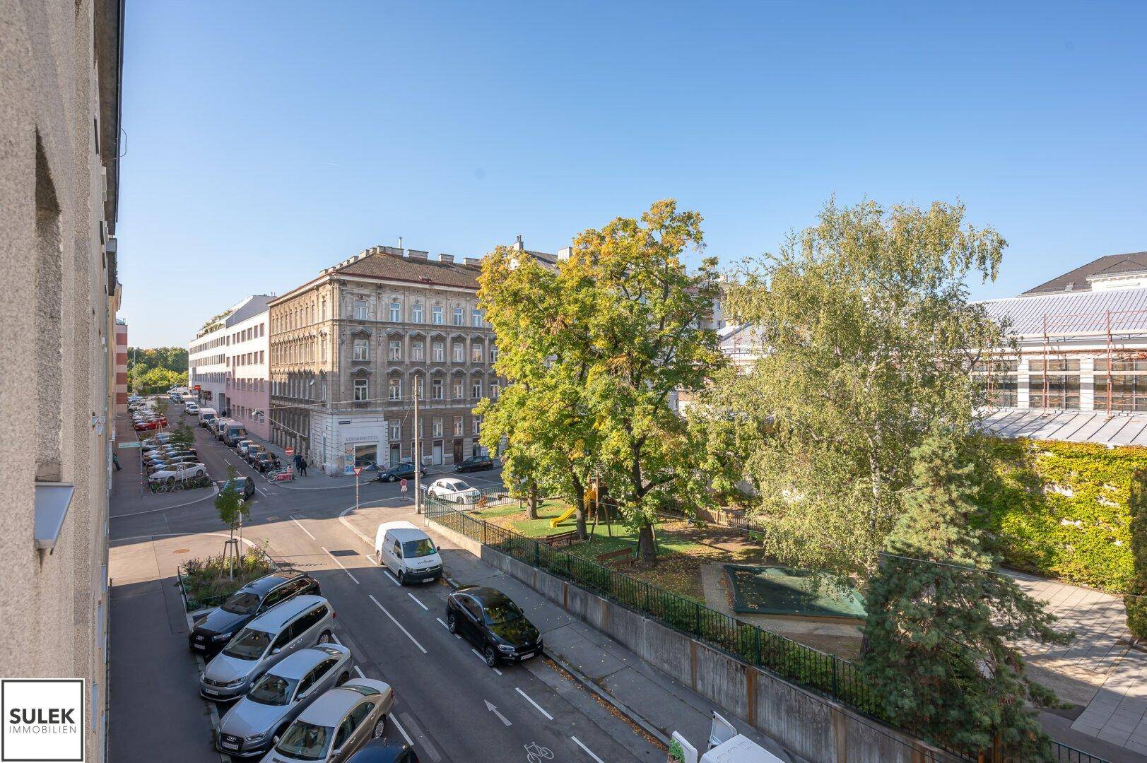 Blick auf die belebte Straße und einen kleinen Park mit Bäumen und parkenden Autos.