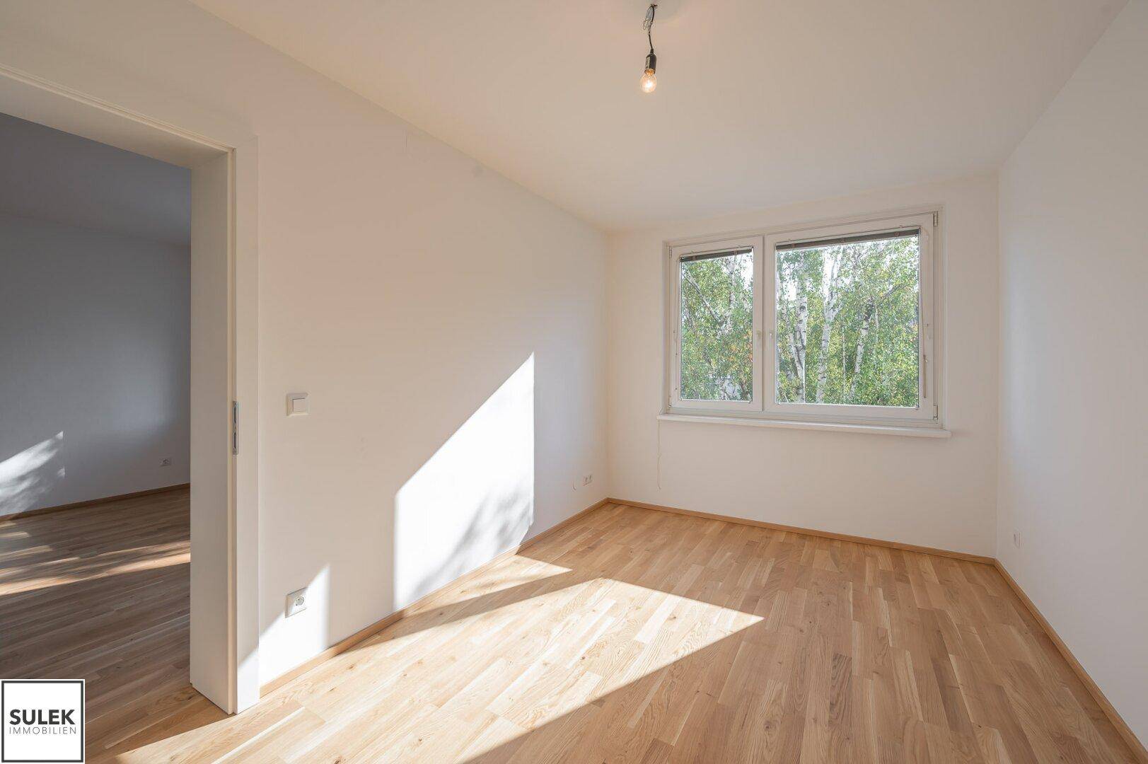 Helles Schlafzimmer mit Parkettboden und einem Fenster, das den Blick ins Grüne freigibt.