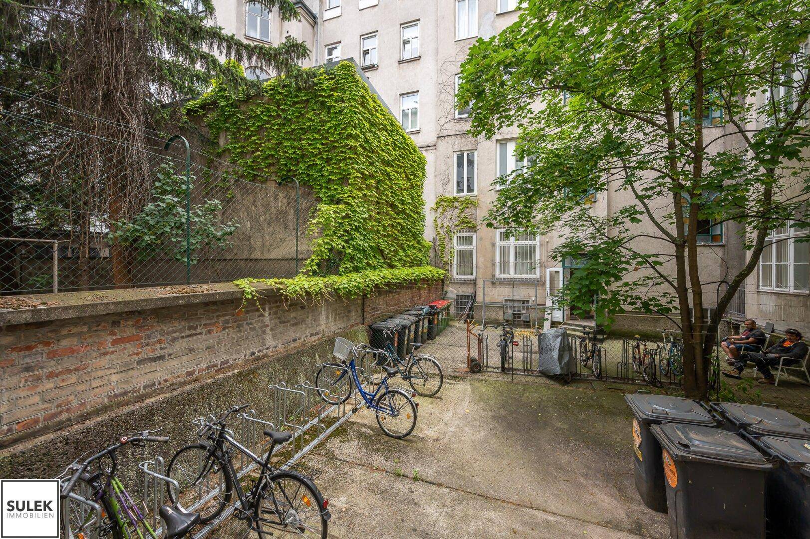 Begrünter Hinterhofbereich eines Mehrfamilienhauses mit Fahrradständern und Mülltonnen.
