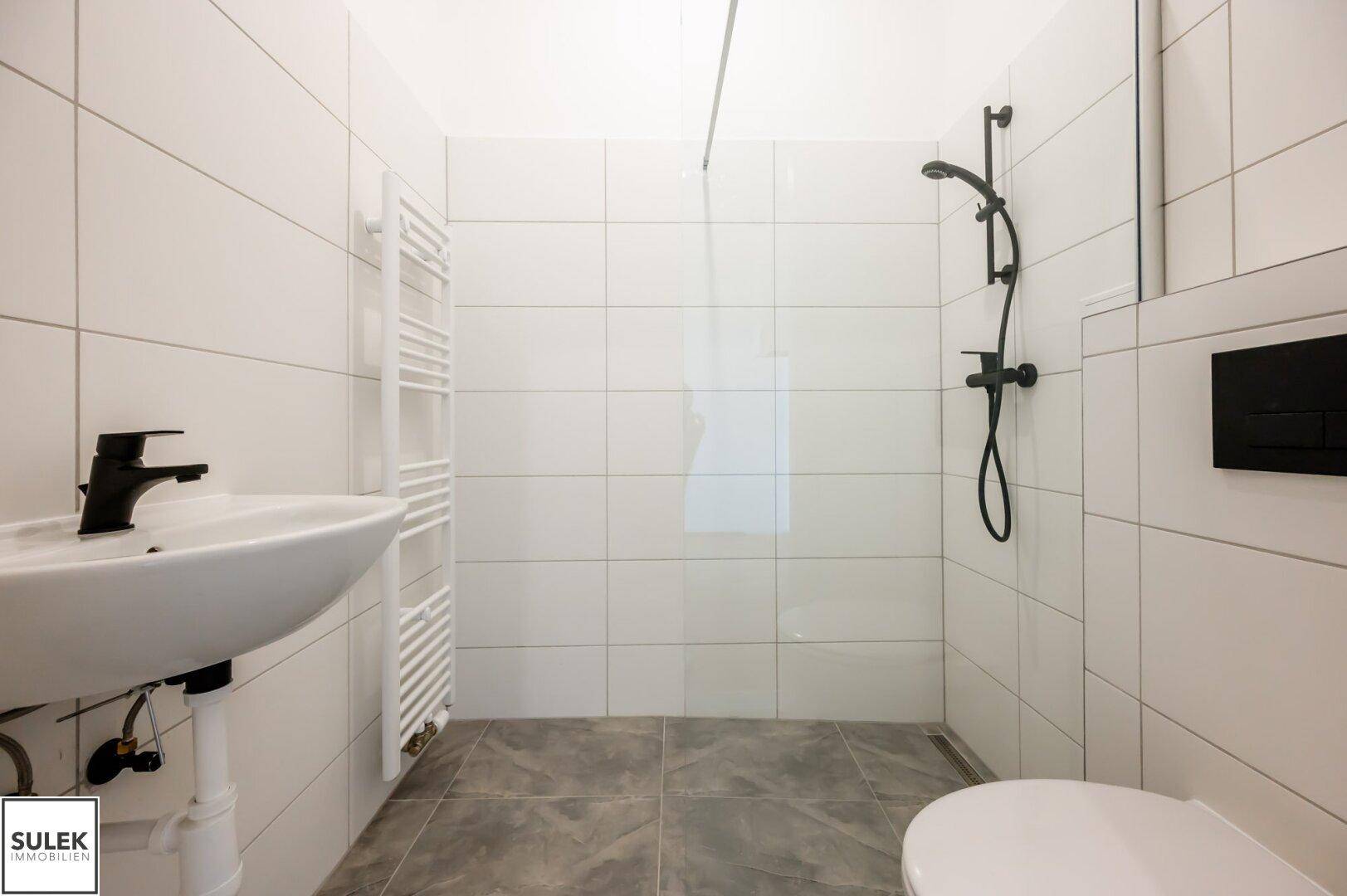 Modernes Badezimmer mit weißen Fliesen, ebenerdiger Dusche, Waschbecken und Handtuchheizkörper.