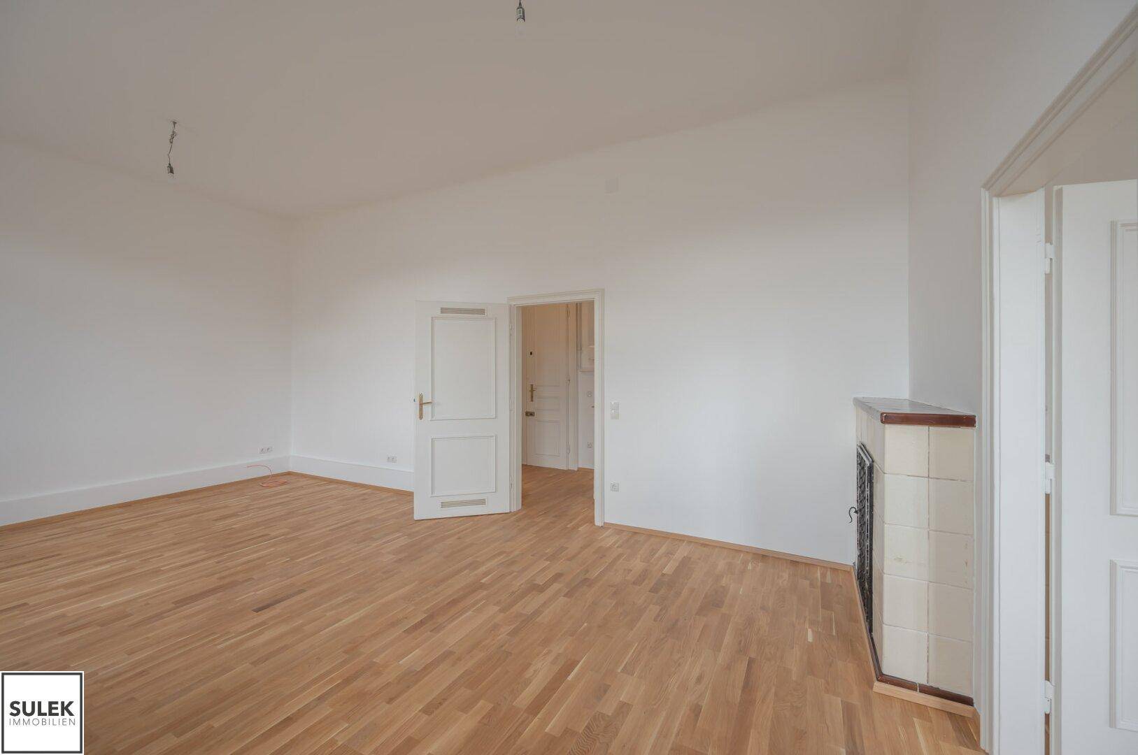 Geräumiger, leerer Raum mit Holzboden und einem traditionellen Kachelofen als Blickfang.