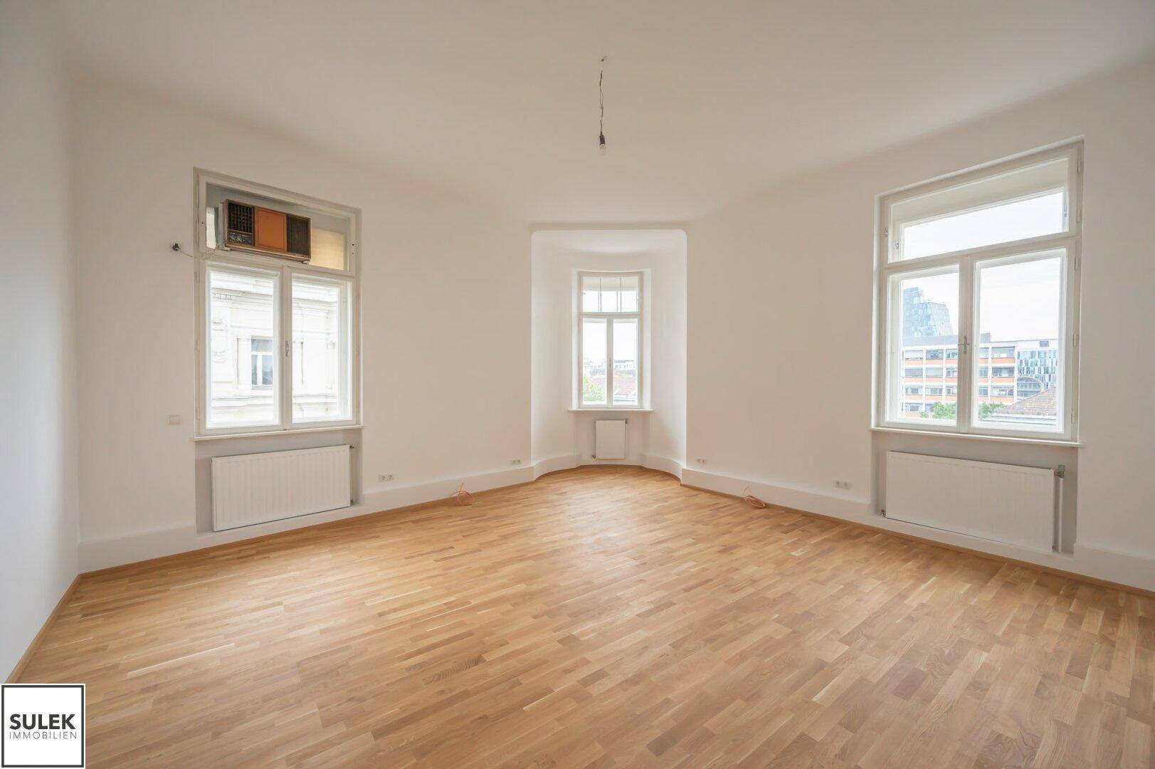 Heller, leerer Raum mit Parkettboden und mehreren Fenstern, die viel Tageslicht hereinlassen.