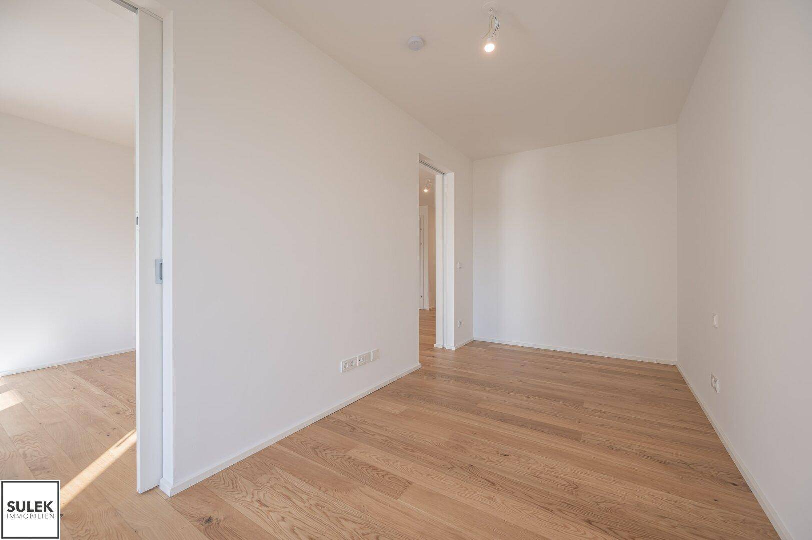 Leeres Zimmer mit hellem Holzboden und Zugang zu einem weiteren Raum.