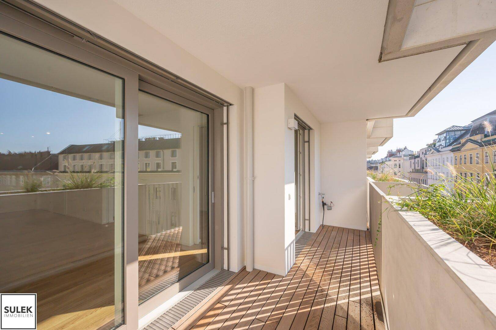 Heller Balkon mit Holzboden und großen Schiebetüren, umgeben von Grünpflanzen.