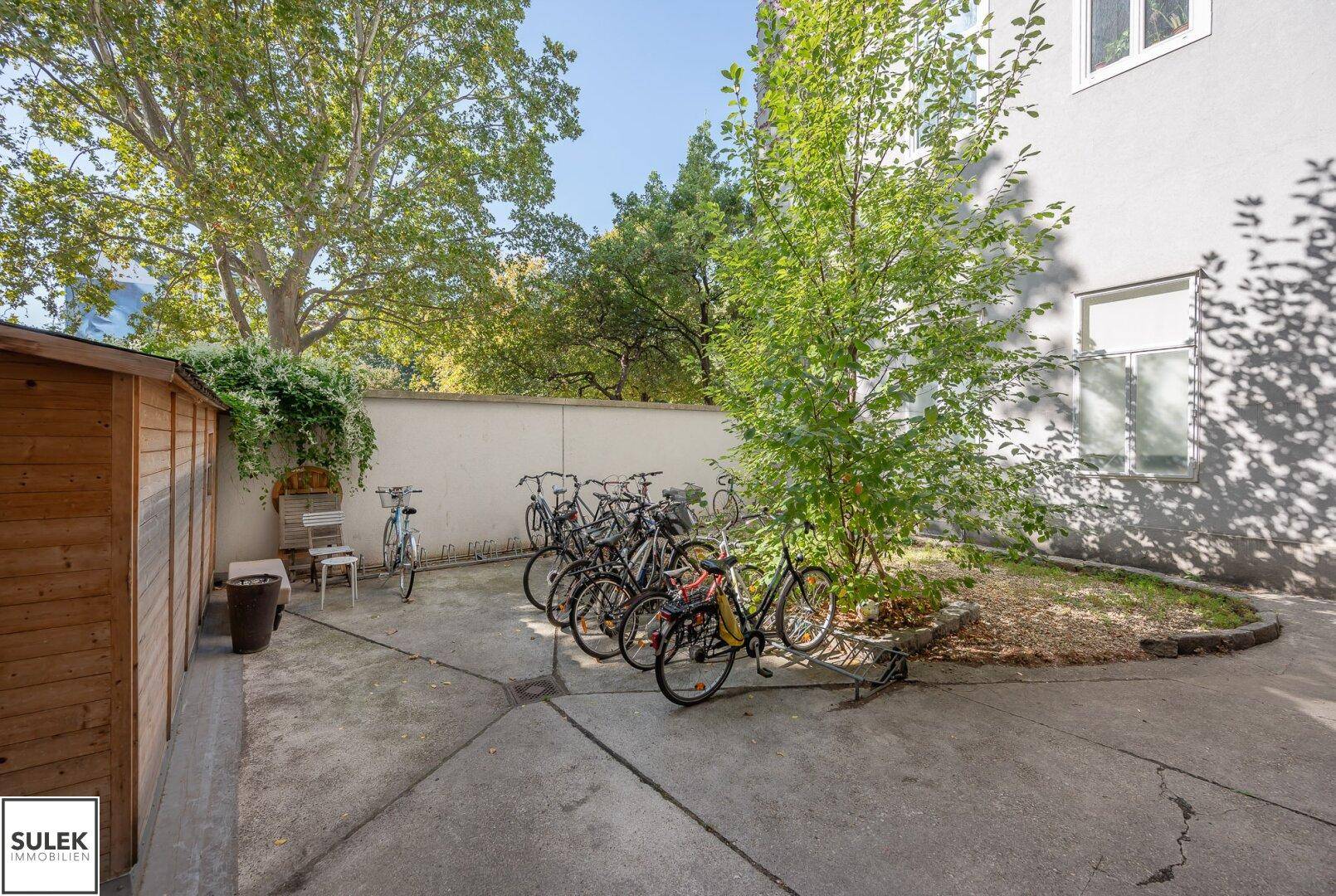 Gemeinschaftlicher Außenbereich mit Fahrradabstellplätzen, Bäumen und einem kleinen Gartenschuppen.