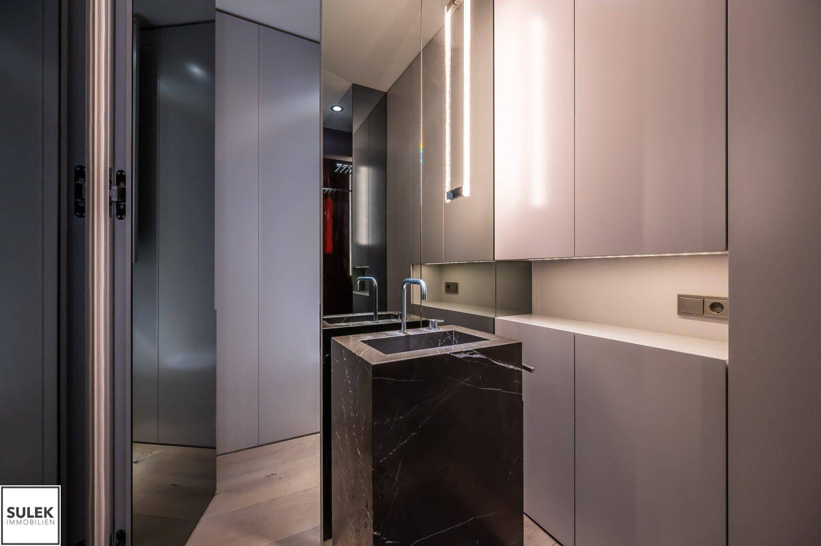 Modernes Badezimmer mit dunklem Marmorwaschbecken, Spiegelflächen und integrierter Beleuchtung.