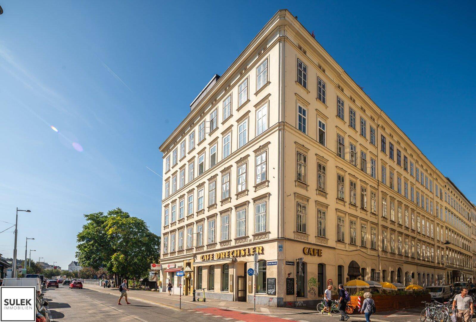 Historisches Eckgebäude mit gelber Fassade, Geschäften im Erdgeschoss und belebter Straße.