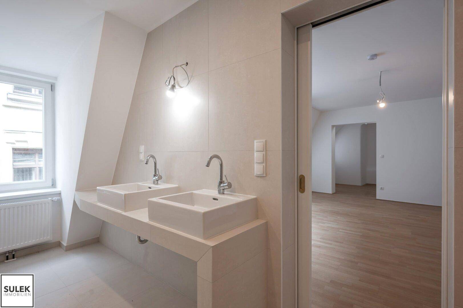 Helles Badezimmer mit Doppelwaschbecken, großem Spiegel und Fenster, das viel Tageslicht hereinlässt.