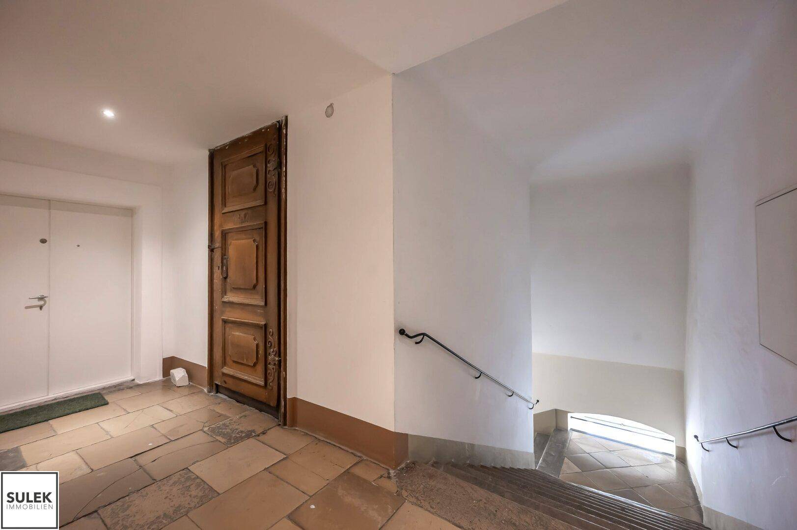 Historischer Flur mit Holztür, Steintreppe und schmiedeeisernem Geländer.