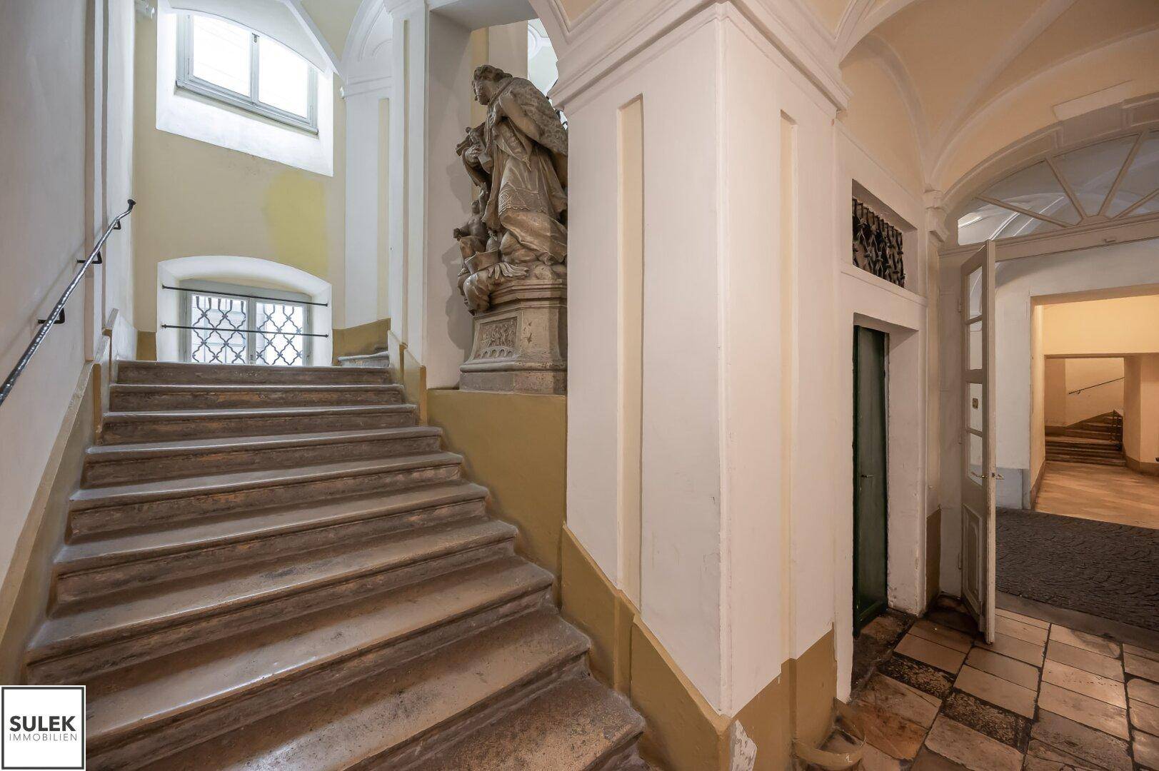 Historisches Treppenhaus mit Steinstufen, einer Statue und gewölbten Fenstern.