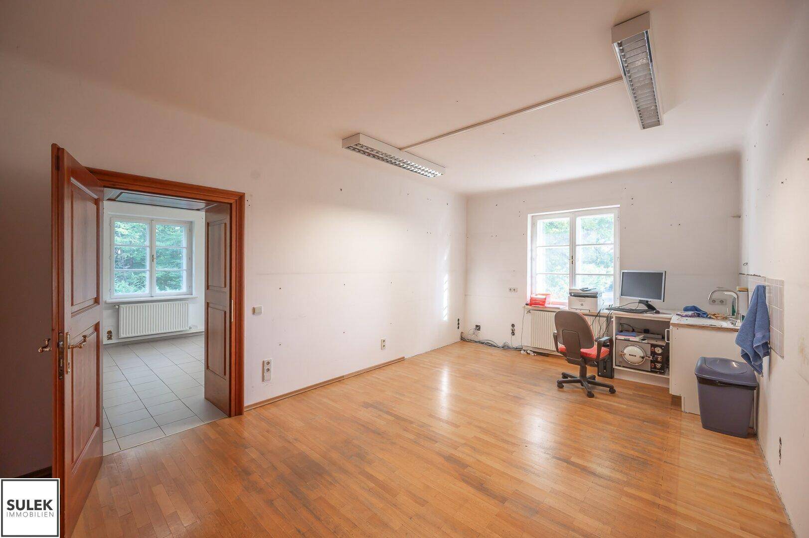 Helles Büro mit Holzfußboden, zwei Fenstern und einfacher Büroeinrichtung, ideal für konzentriertes Arbeiten.