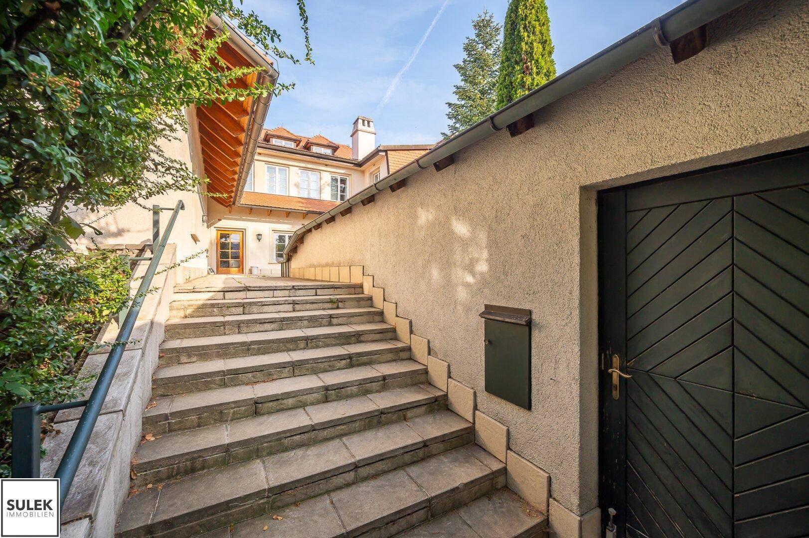 Steintreppe führt zum Eingangsbereich des Hauses, umgeben von einer Mauer und üppigen Grünflächen.