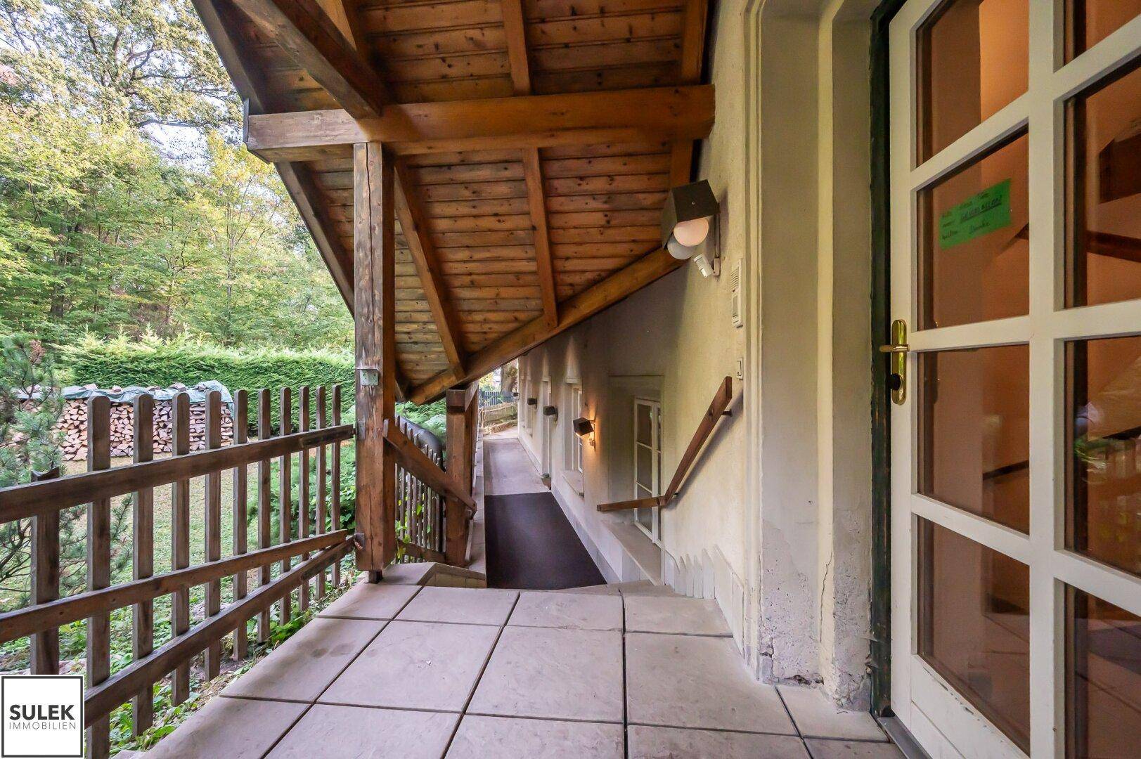 Überdachter Eingangsbereich mit Fliesenboden und Holzgeländer, umgeben von viel Grün und natürlichem Licht.