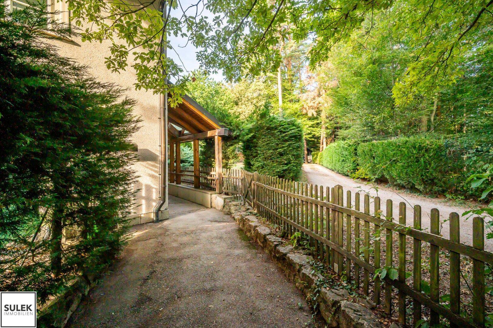 Schmaler Weg führt durch grüne Vegetation zum Haus, umgeben von Bäumen und einem Holzzaun.