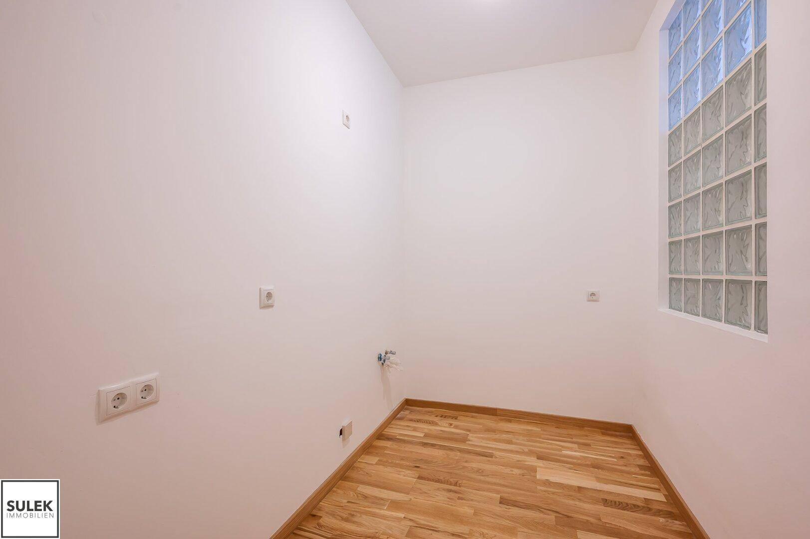 Kleiner Raum mit Parkettboden, weißen Wänden und einem Fenster aus Glasbausteinen.