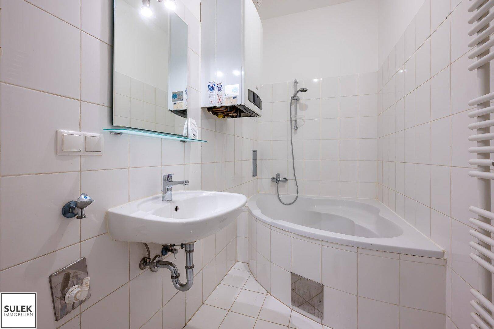 Funktionales Badezimmer mit weißen Fliesen, Eckbadewanne und Waschbecken.