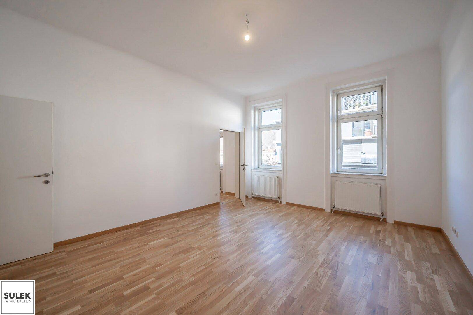 Geräumiges Zimmer mit Parkettboden, weißen Wänden und zwei großen Fenstern.