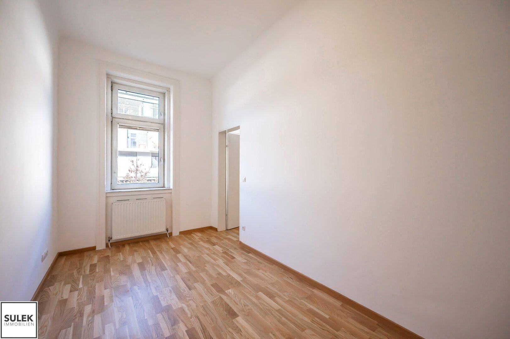 Helles Zimmer mit Parkettboden, weißen Wänden und einem Fenster mit Ausblick.