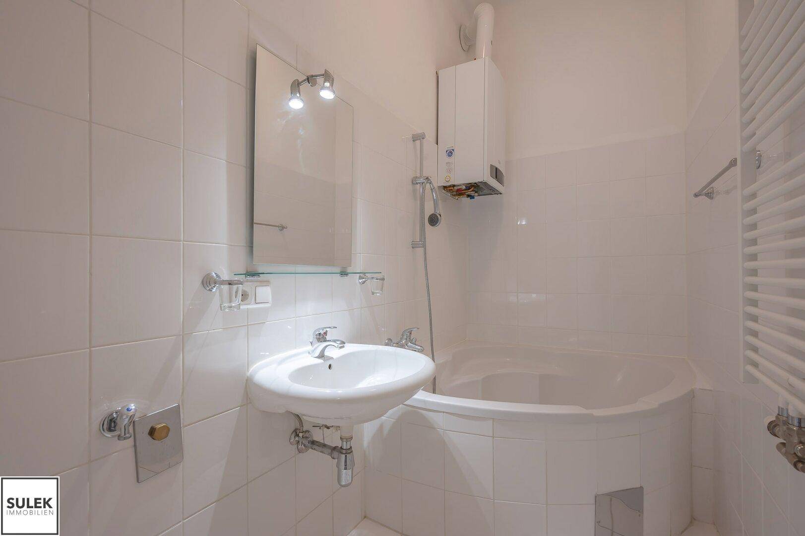 Helles Badezimmer mit weißer Eckbadewanne, Waschbecken und Wandfliesen, inklusive Warmwasserboiler.