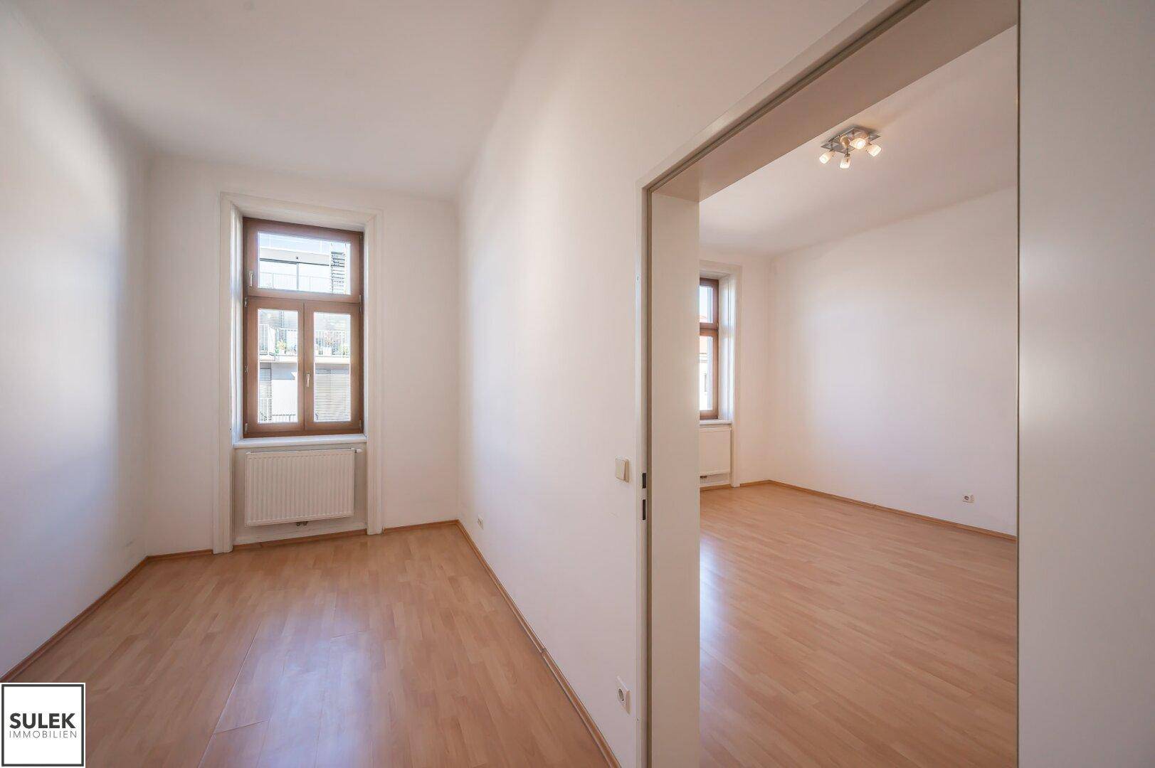 Flur mit Parkettboden, der zu einem weiteren hellen Zimmer mit Fenstern führt.