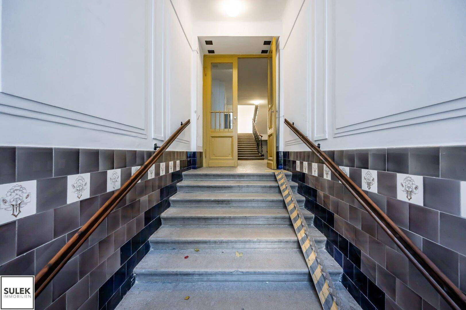 Eingangsbereich eines Altbaus mit Treppe, dekorativen Fliesen und einer Rollstuhlrampe.