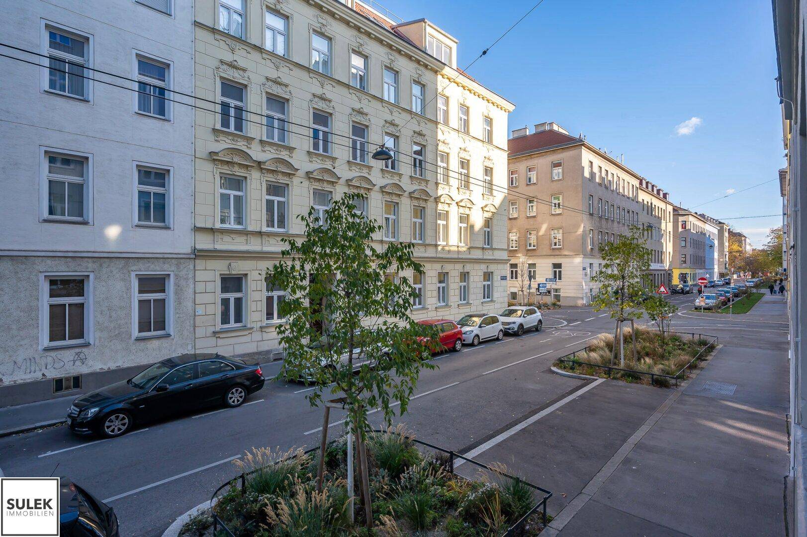 Ruhige Wohnstraße mit parkenden Autos und begrünten Baumbeeten an einem sonnigen Tag.