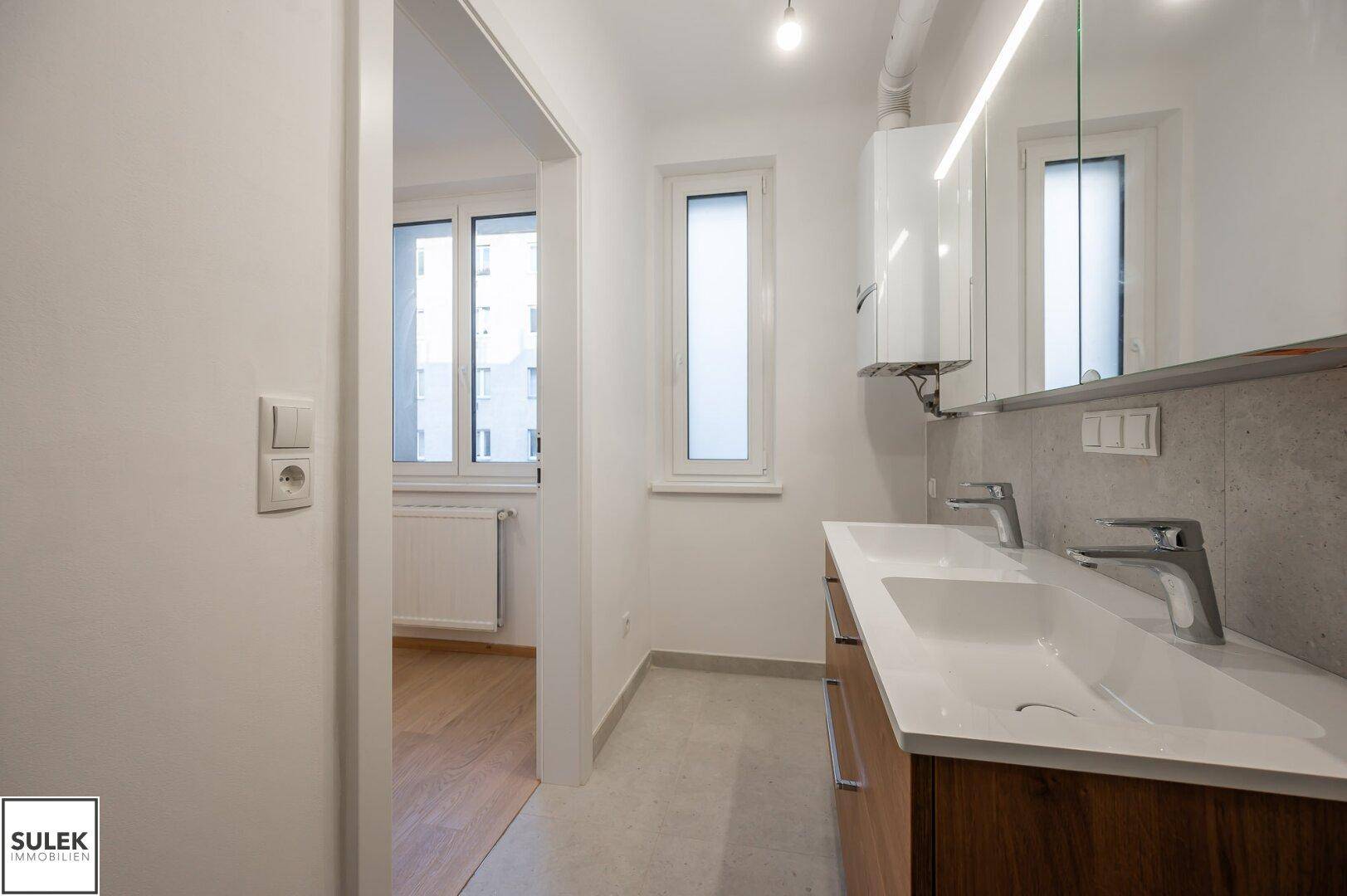 Modernes Badezimmer mit Doppelwaschbecken, Fenster und Blick in ein angrenzendes Zimmer.