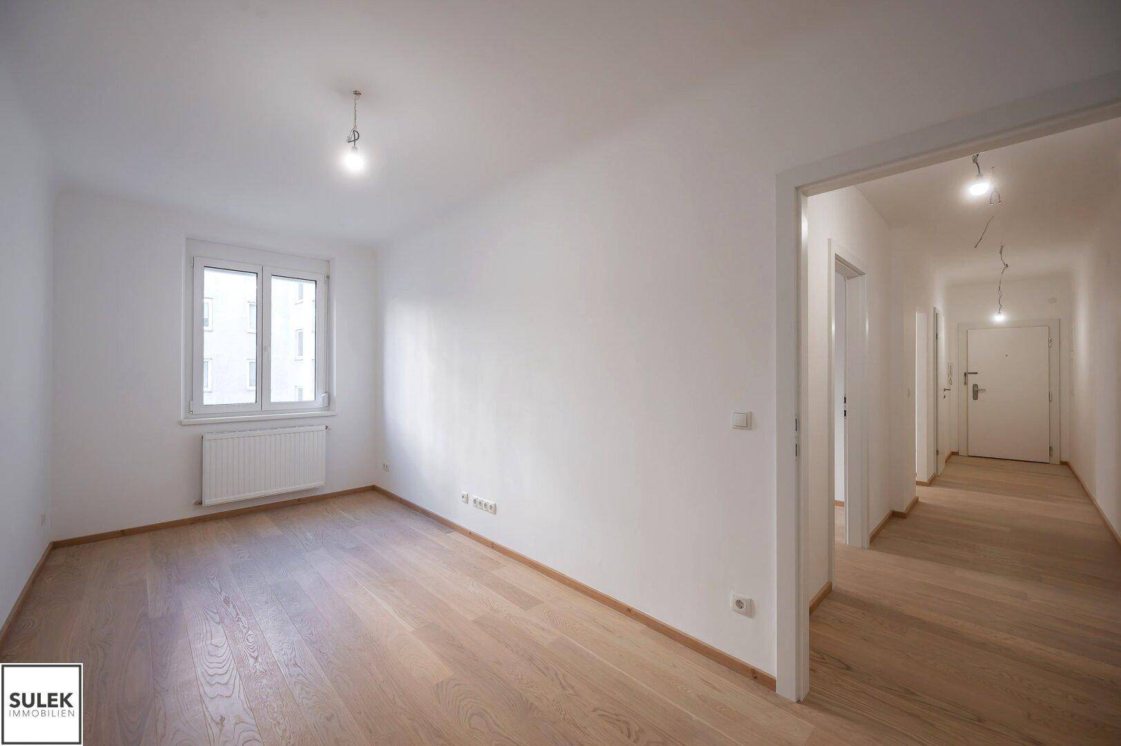 Geräumiges Zimmer mit hellem Holzboden und Blick in den angrenzenden Flur, ideal als Schlafzimmer.