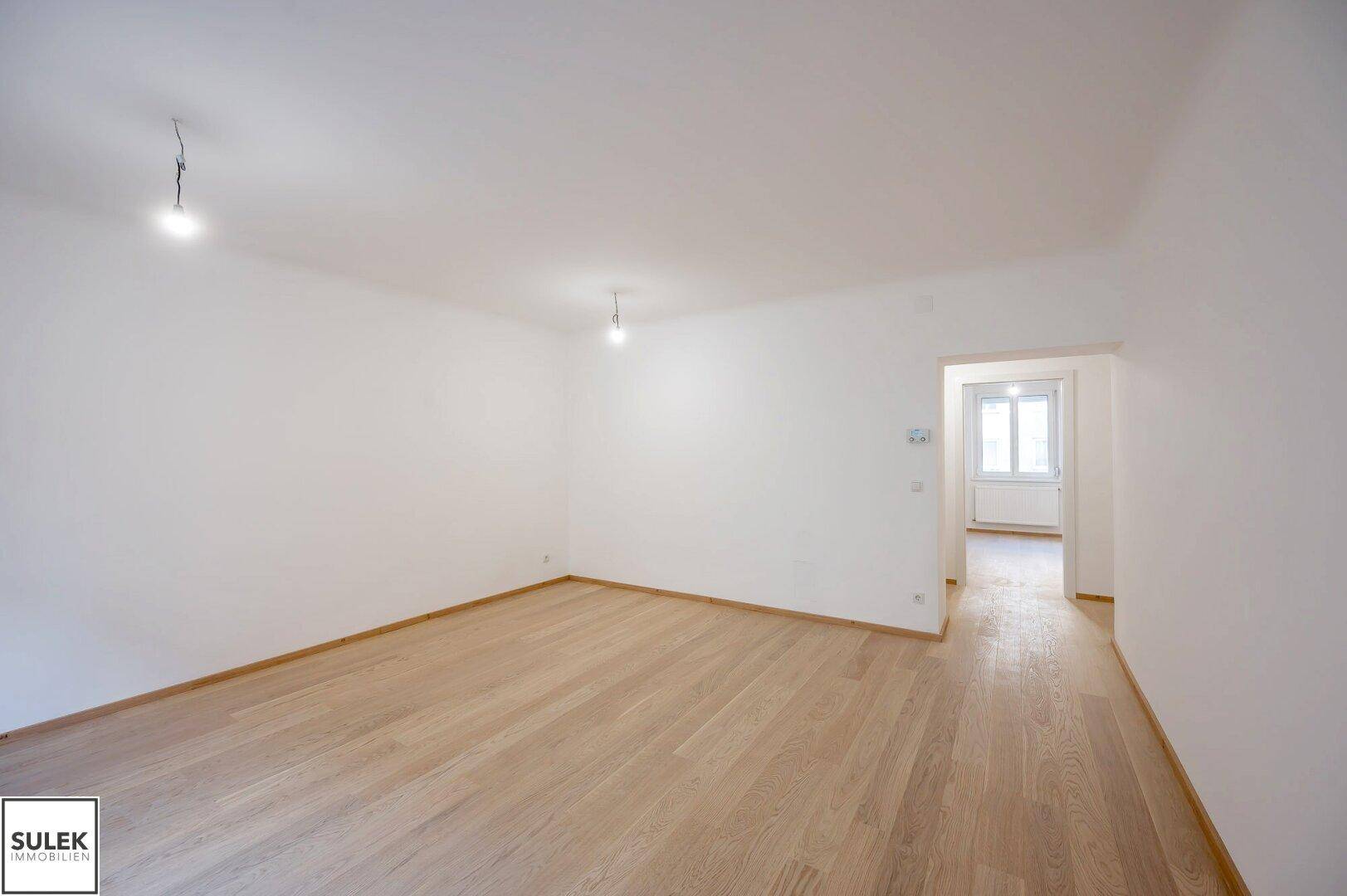 Helles Zimmer mit Holzboden und offenem Übergang zu einem weiteren Bereich der Wohnung.