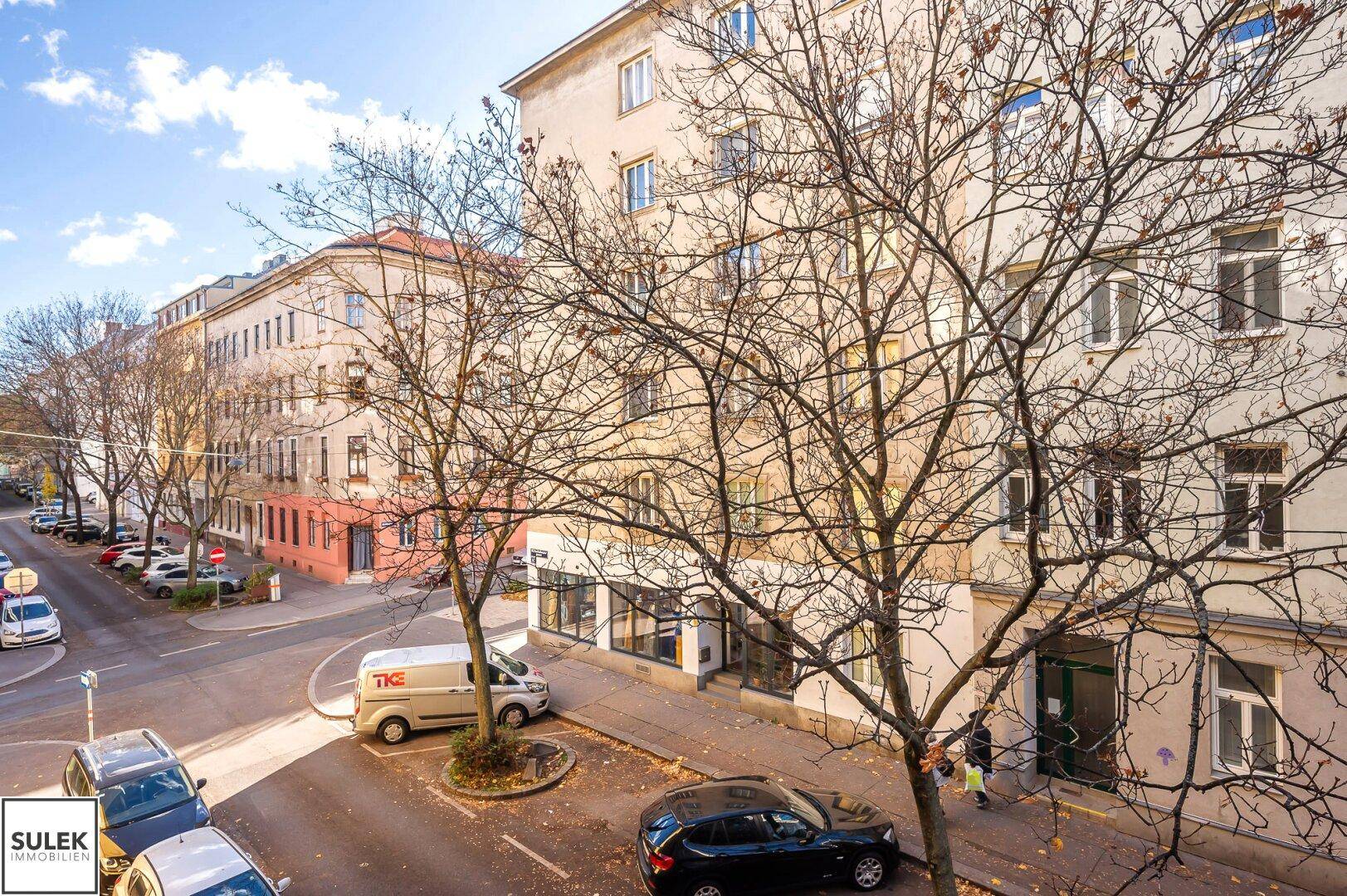 Blick auf eine belebte Straße mit parkenden Autos und umliegenden Wohngebäuden unter blauem Himmel.