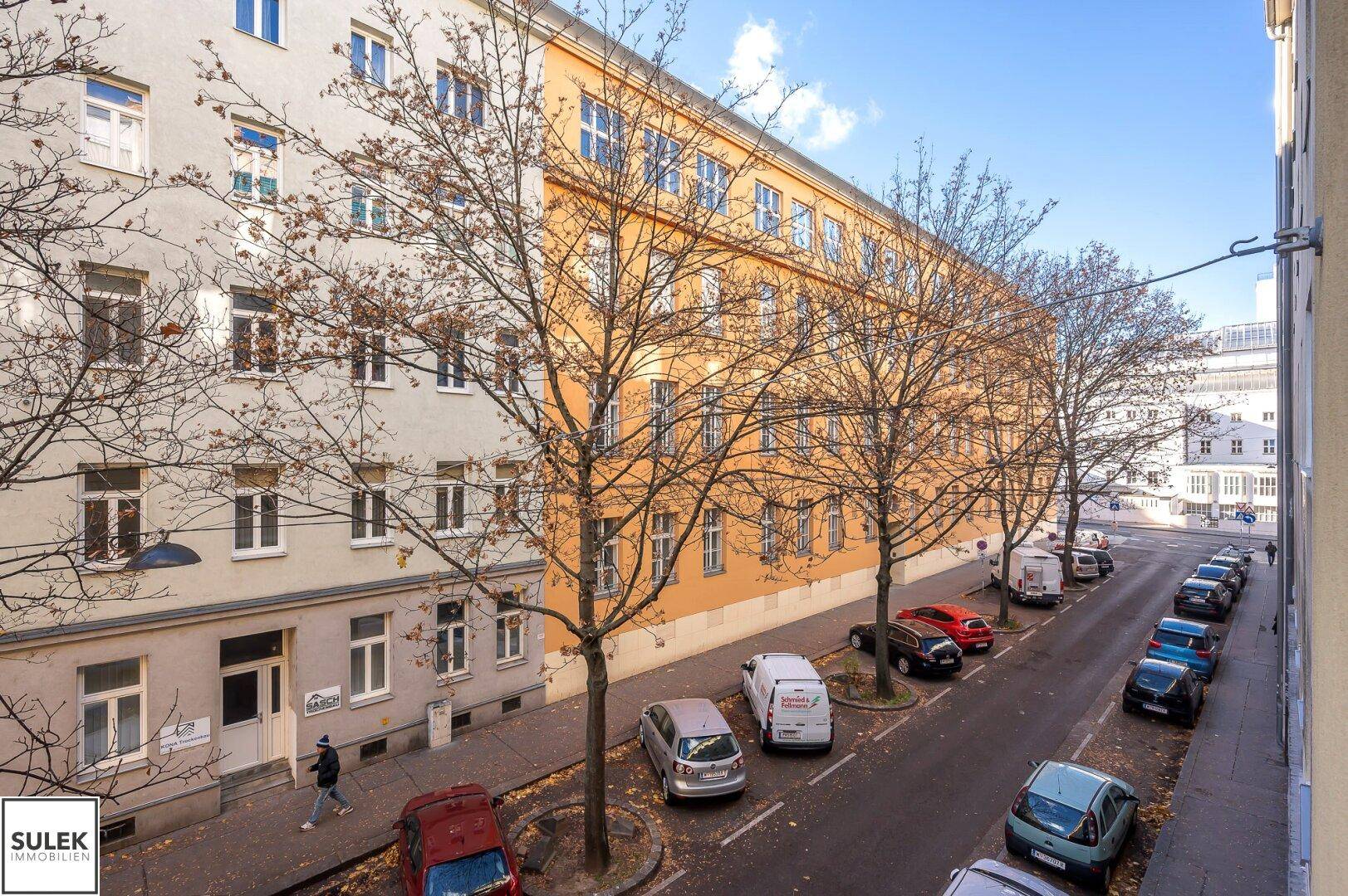Straßenansicht mit parkenden Fahrzeugen und einer Reihe von Wohngebäuden unter blauem Himmel.