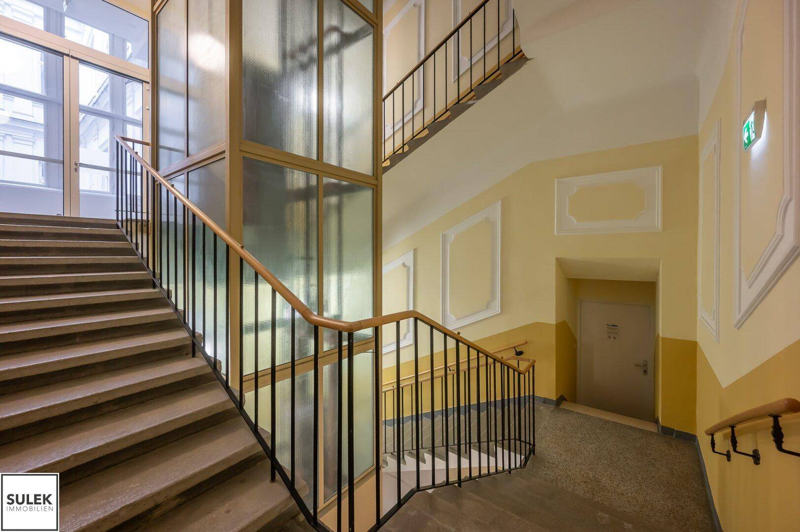 Elegantes Treppenhaus mit modernem Aufzug, Marmorboden und großen Fenstern, die viel Licht hereinlassen.