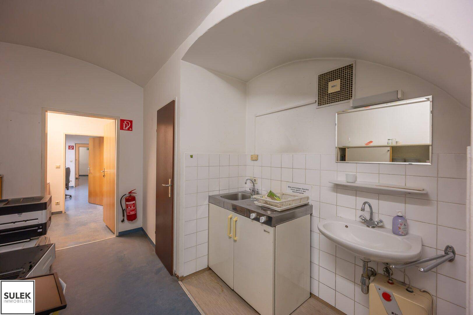 Kleine Küchenzeile mit Spülbecken, Fliesenwand und einfacher Ausstattung, angrenzend an einen Flur.