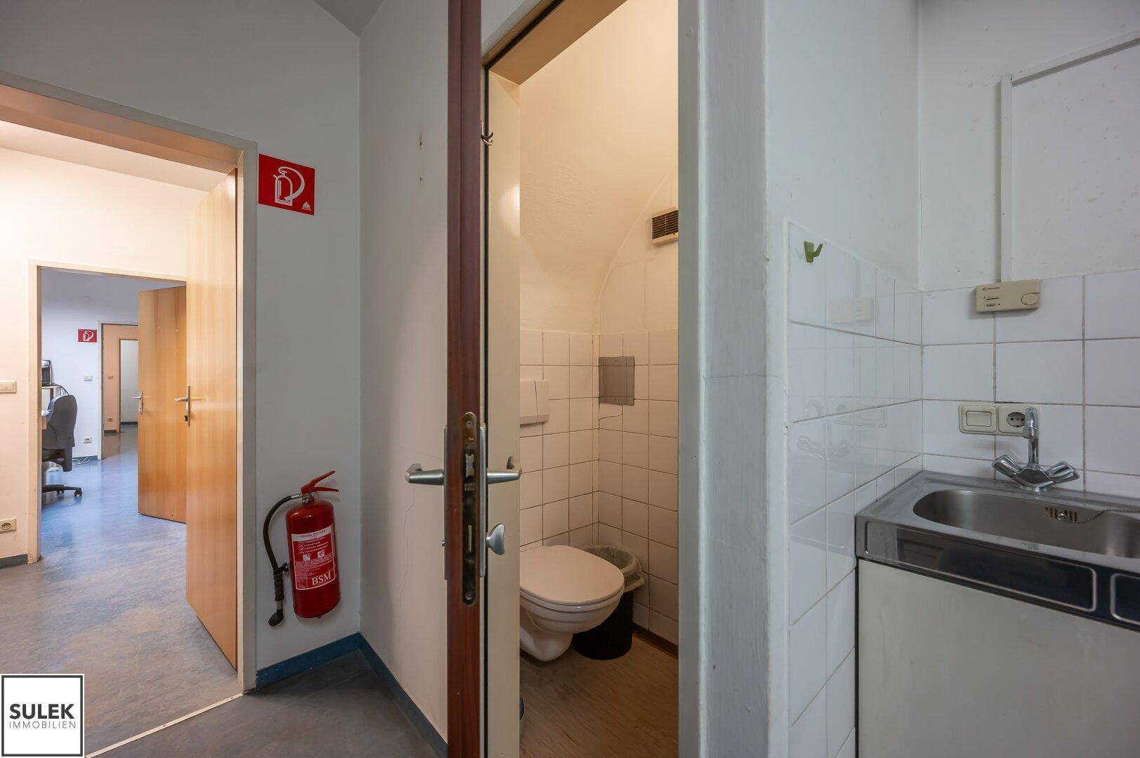 Einfache Toilette mit Fliesen an den Wänden und einem kleinen Waschbecken, zugänglich vom Flur.
