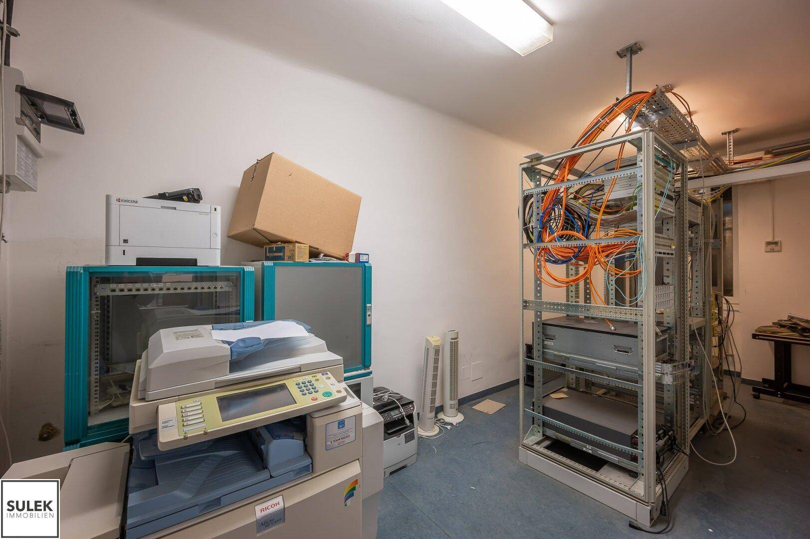 Technikraum mit Serverracks, Druckern und Verkabelung, ausgestattet für IT-Infrastruktur.