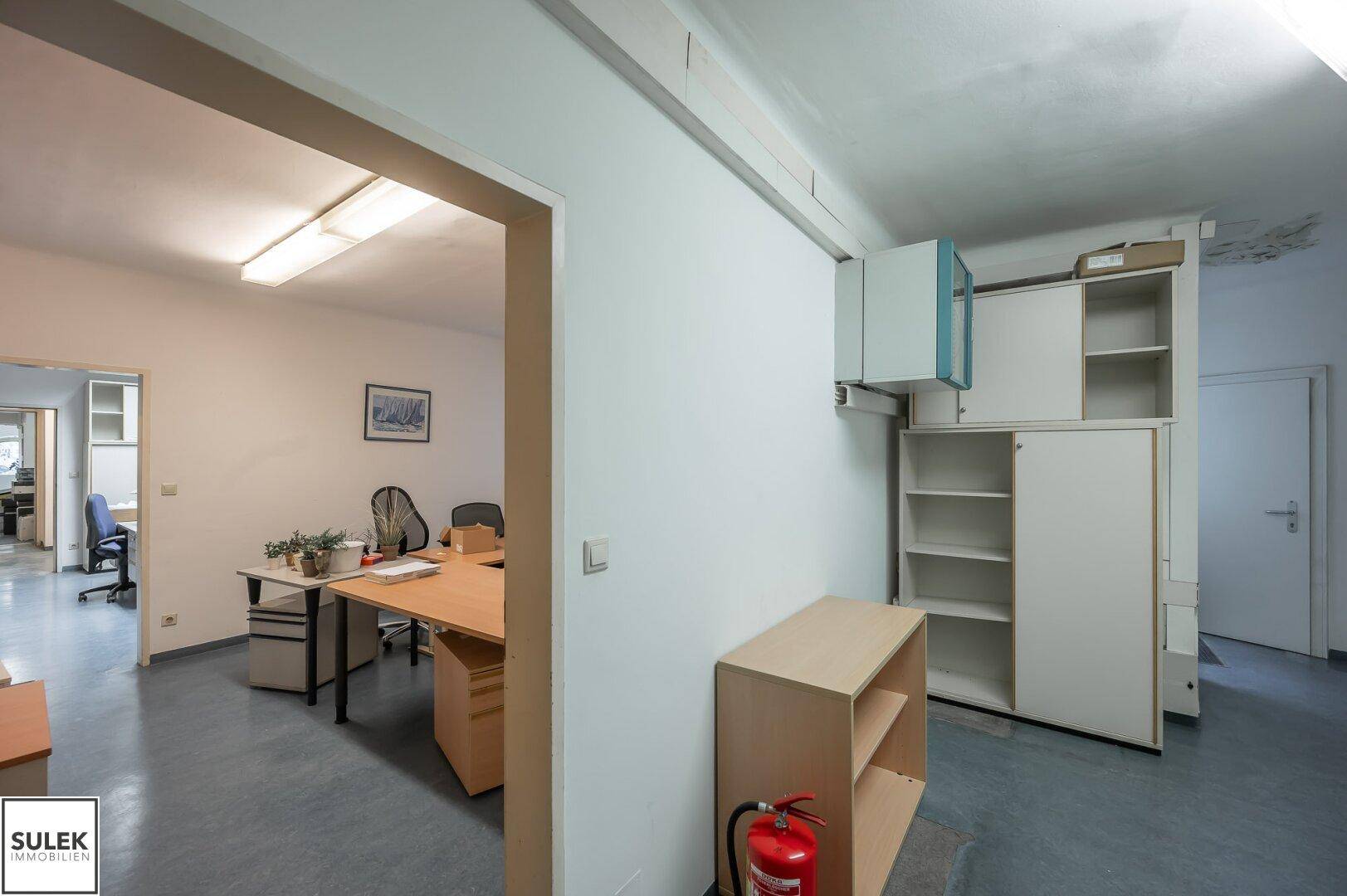 Bürobereich mit Schreibtischen und Regalen, verbunden mit einem Flur, der zu weiteren Räumen führt.