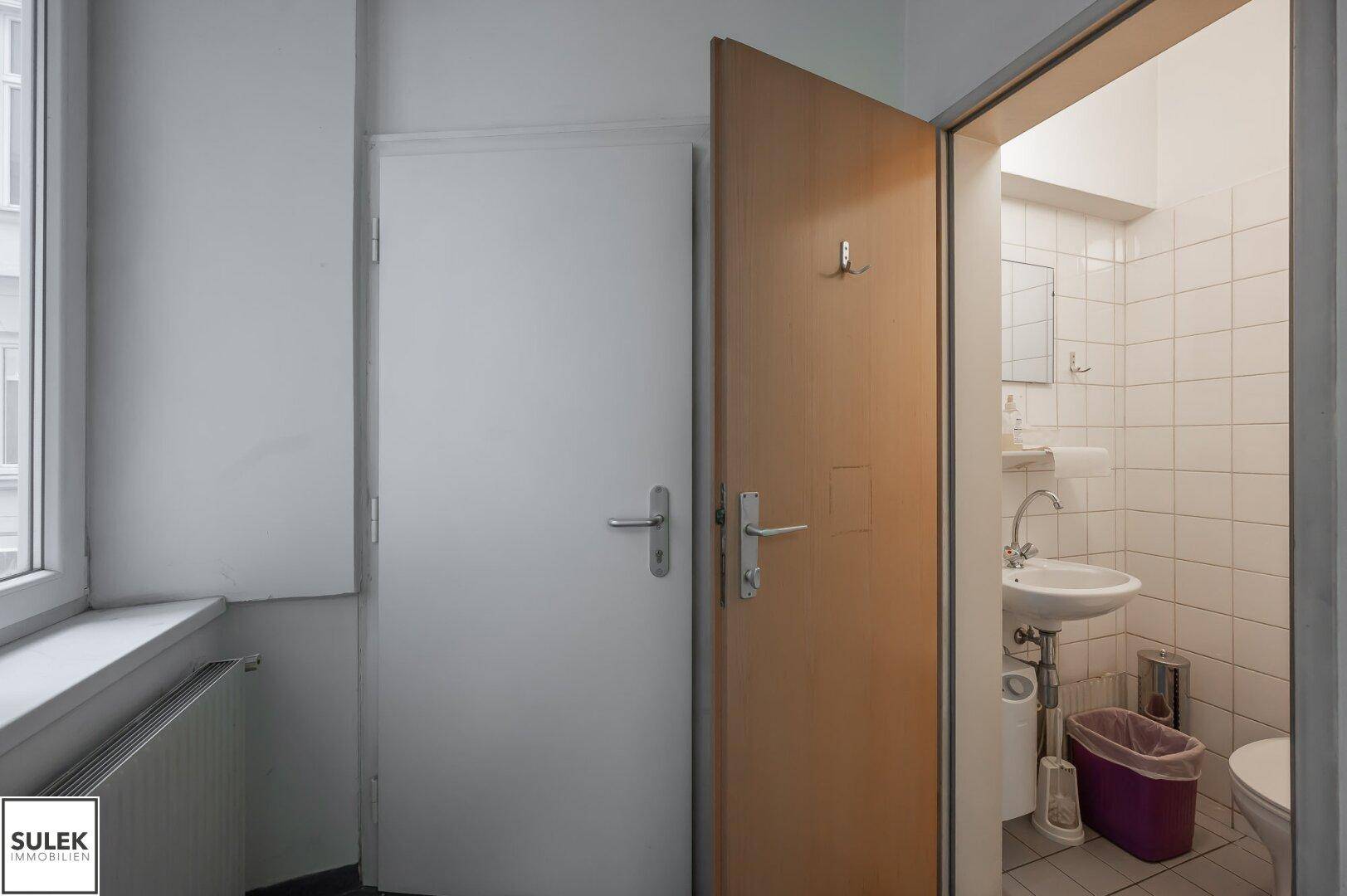 Einfache Toilette mit Fliesen an den Wänden und einem kleinen Waschbecken, zugänglich vom Flur.