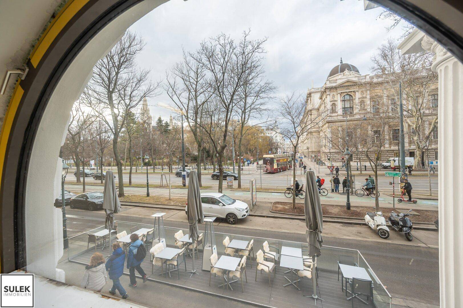Blick aus einem gewölbten Fenster auf eine belebte Stadtstraße mit Gebäuden und Passanten.
