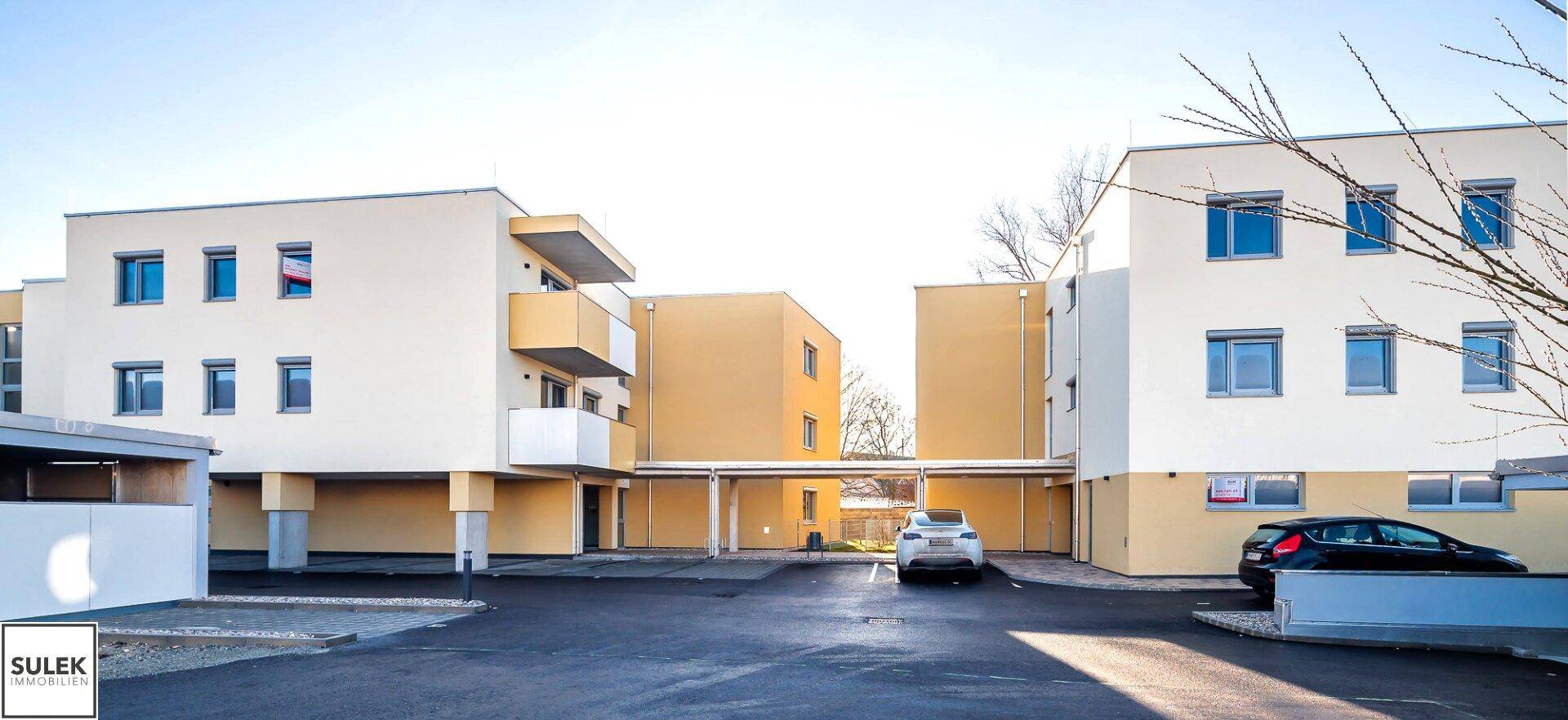 Mehrere moderne Wohngebäude mit hellen Fassaden und einem offenen Parkplatzbereich unter klarem Himmel.