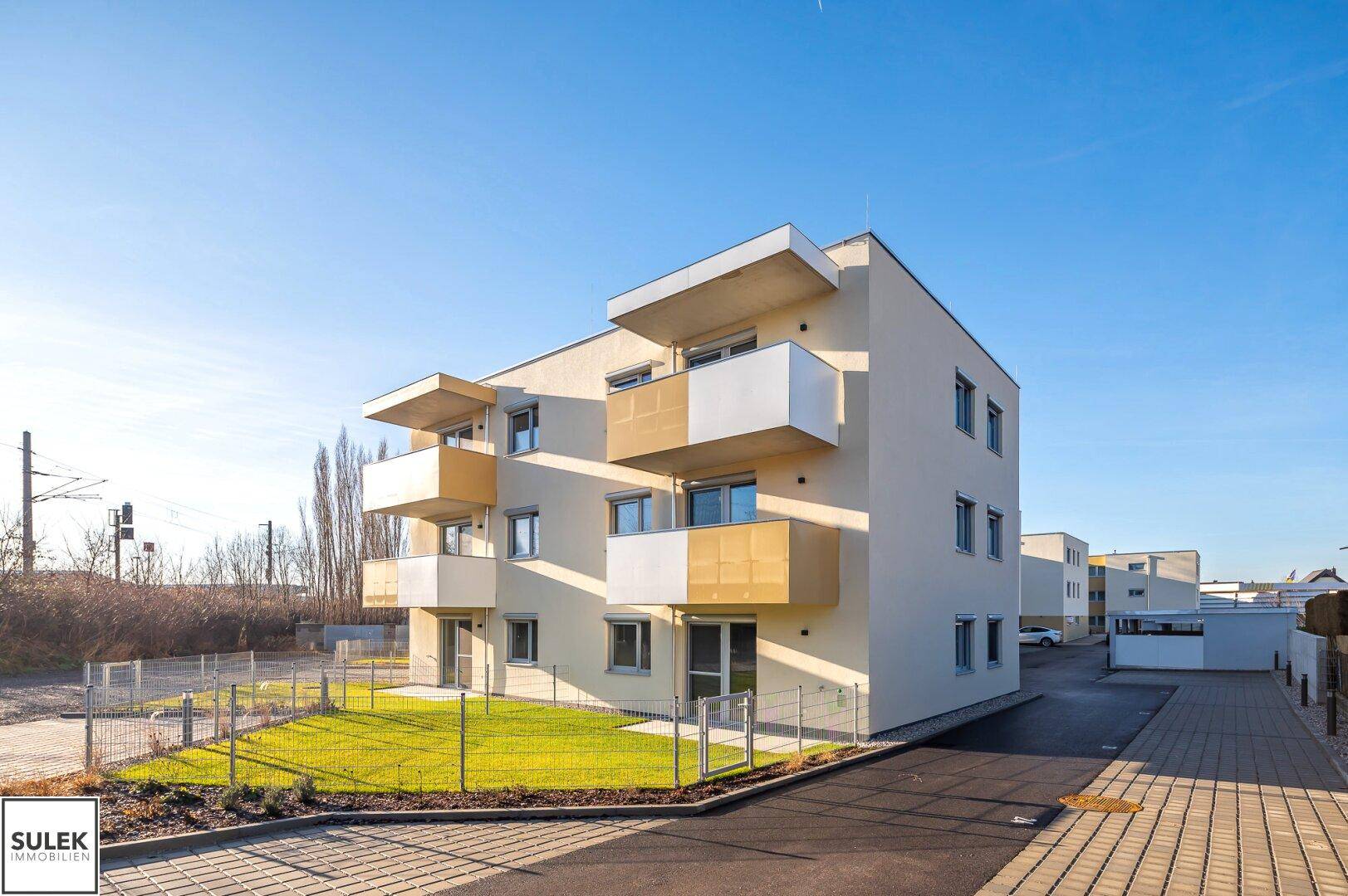 Modernes Mehrfamilienhaus mit gelber Fassade, Balkonen und umzäunten Rasenflächen.