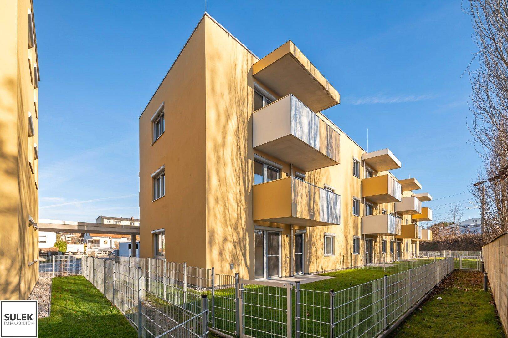 Modernes Mehrfamilienhaus mit gelber Fassade, Balkonen und umzäunten Rasenflächen.
