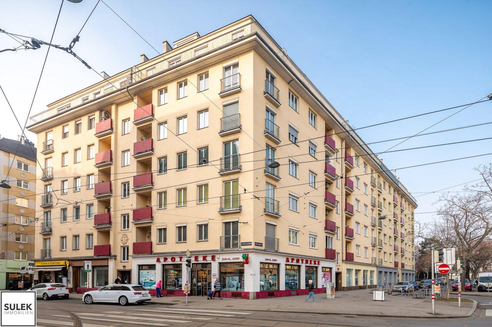 Großes Eckgebäude mit mehreren Etagen, Balkonen und Geschäften im Erdgeschoss an einer Stadtkreuzung.
