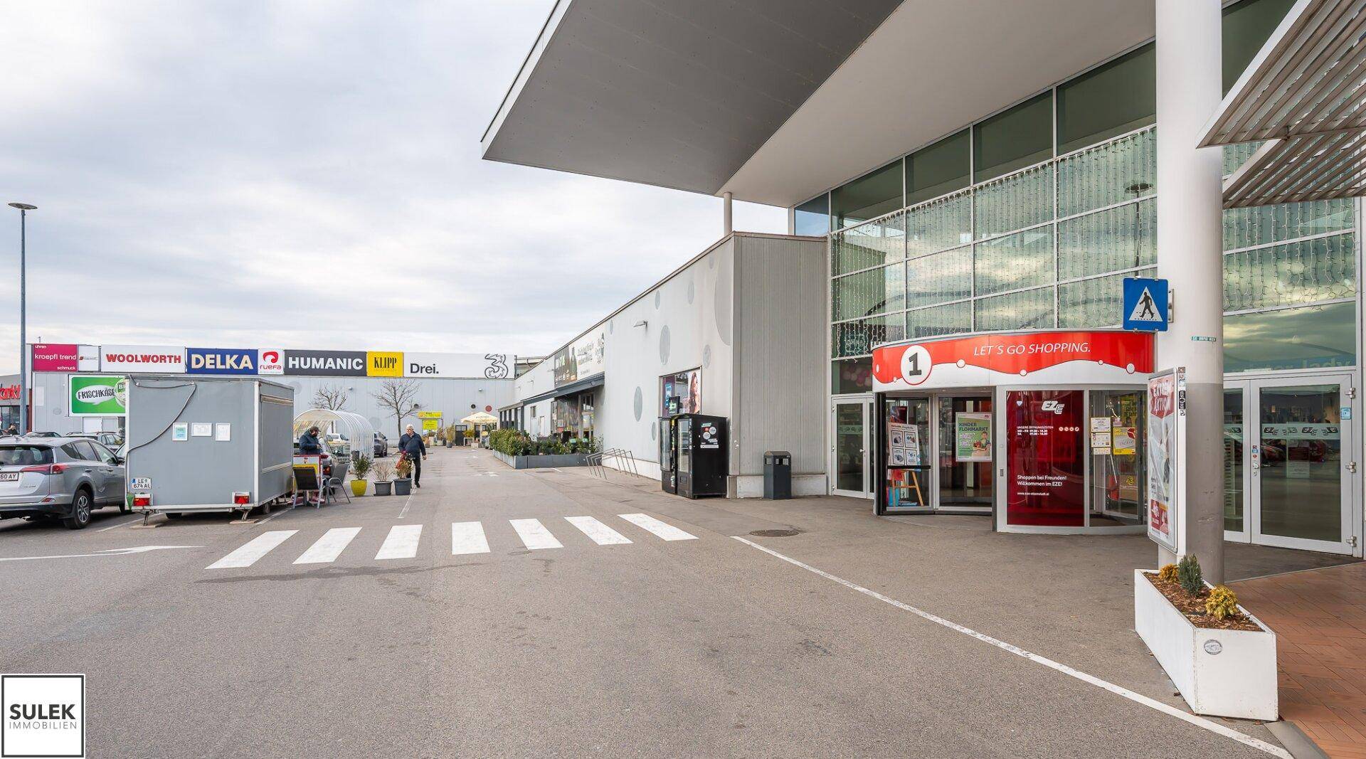 Einkaufszentrum mit verschiedenen Geschäften und Parkplätzen unter bewölktem Himmel.
