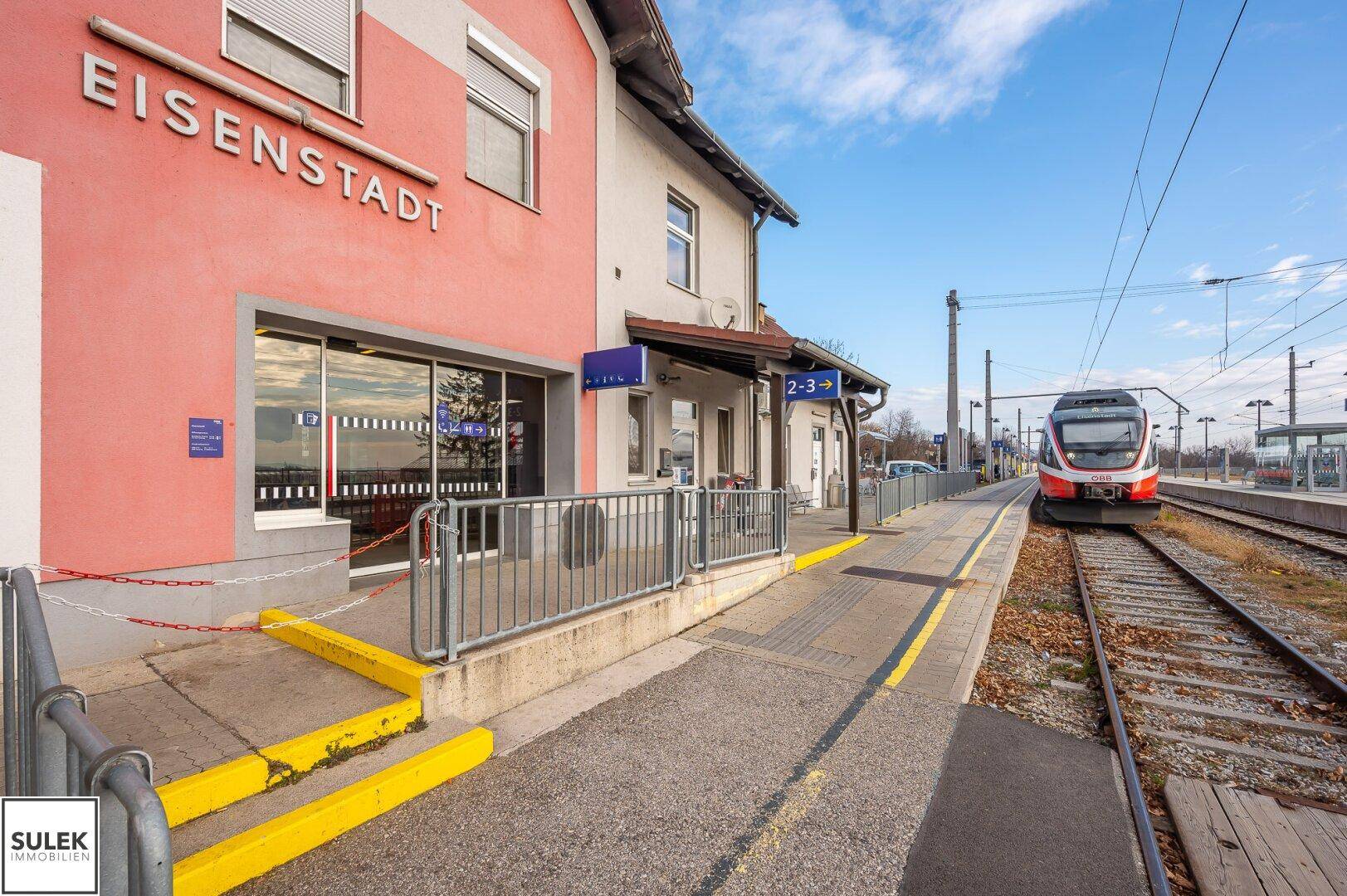 Bahnhofsgebäude in Eisenstadt mit Bahnsteig und einfahrendem Zug unter blauem Himmel.