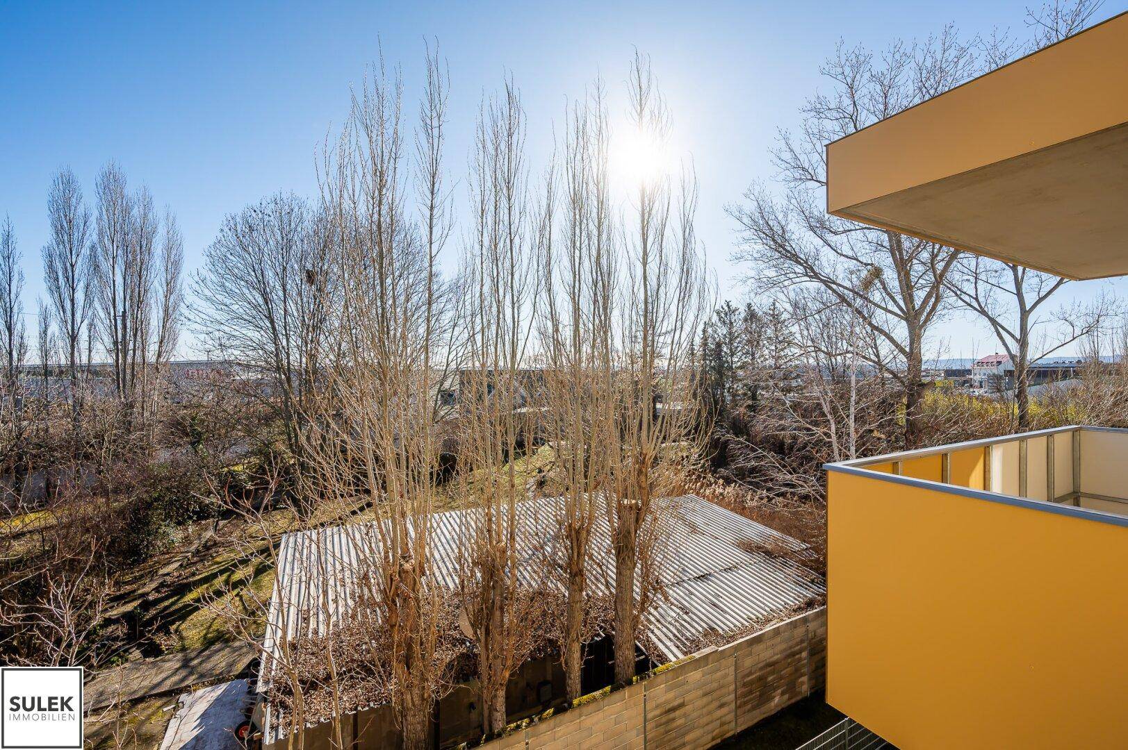 Sonniger Ausblick von einem Balkon auf die grüne Umgebung und weitere Gebäude.