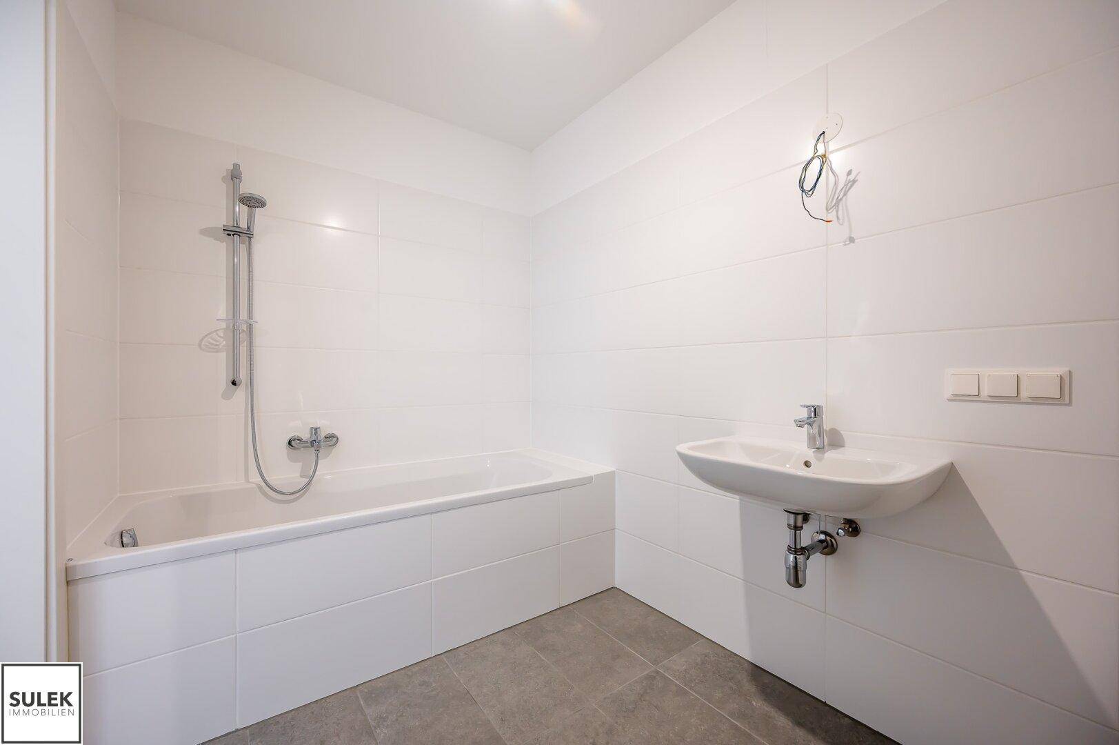 Helles Badezimmer mit Badewanne, Duscharmatur und Waschbecken, ausgestattet mit weißen Fliesen.