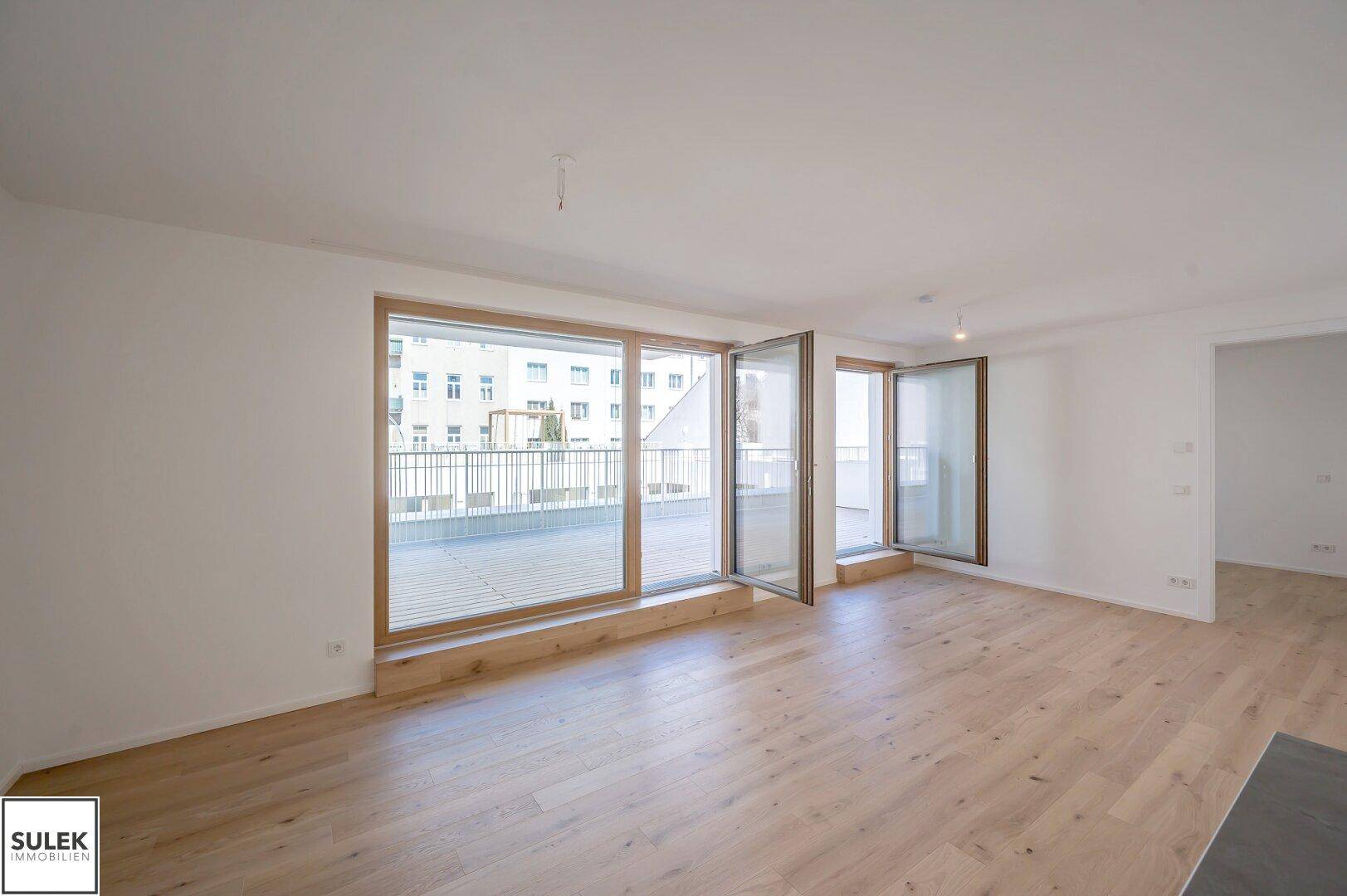 Heller, leerer Wohnraum mit Holzboden und großen Fenstern, die Zugang zu einer Terrasse bieten.