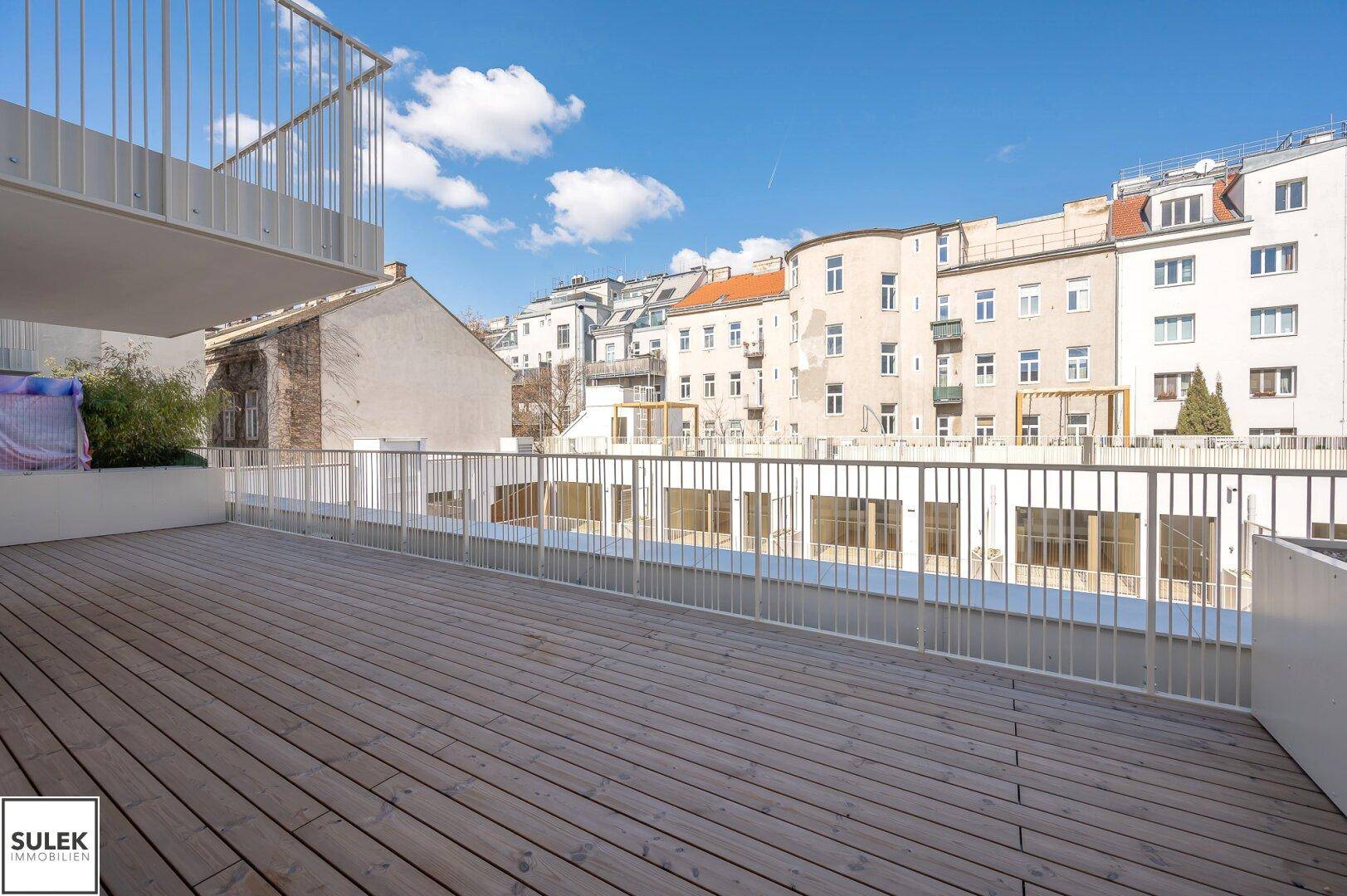 Geräumige Holzterrasse mit Metallgeländer und Blick auf die umliegenden Stadtgebäude unter blauem Himmel.